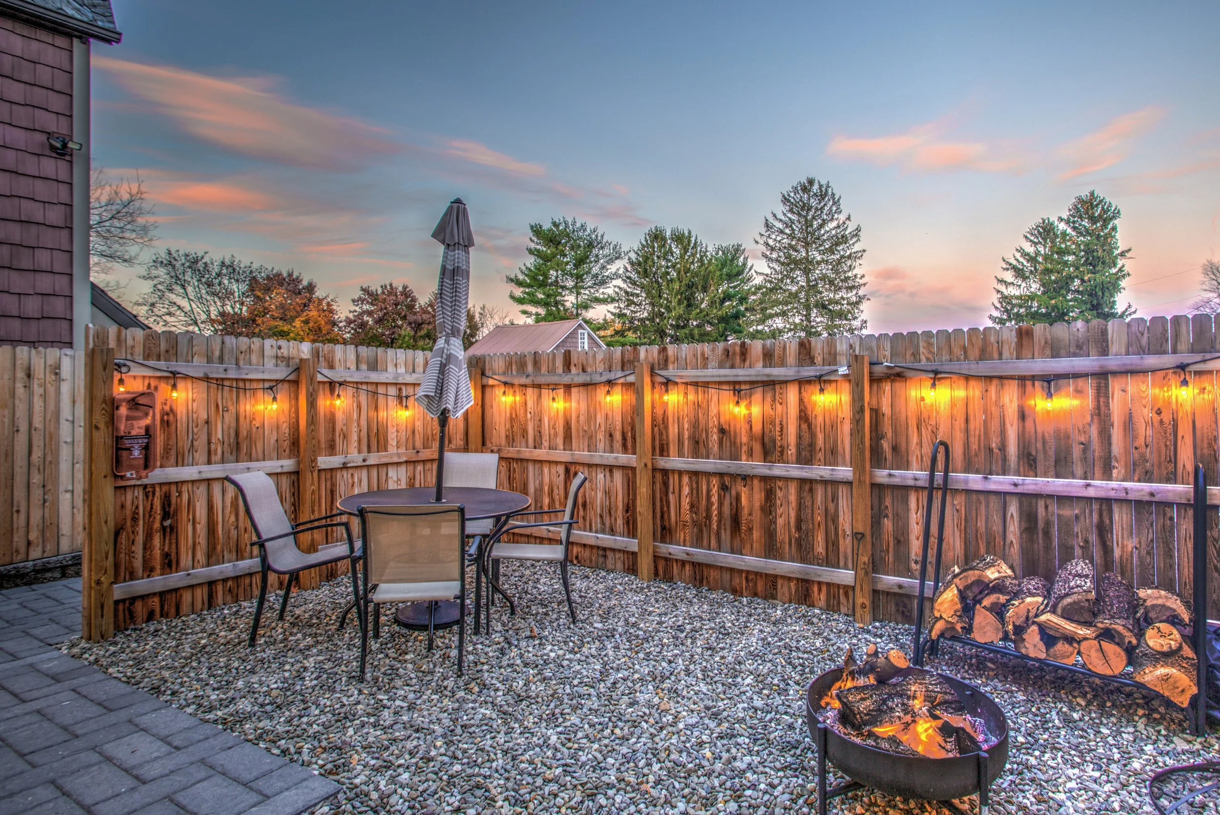A backyard patio at dusk with a wooden fence decorated with string lights, a table with chairs and a closed umbrella, and a fire pit with burning logs. Trees and houses are visible in the background against a colorful sky.