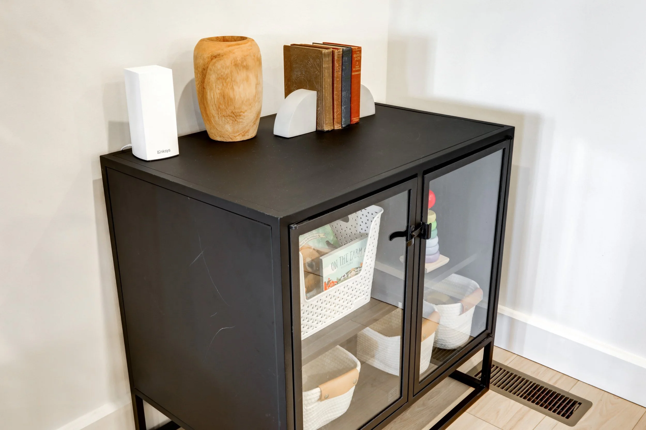 A black glass-front cabinet with decorative objects on top, including a large wooden vase, books, and white bookends. Inside, visible through the glass, are baskets and books.