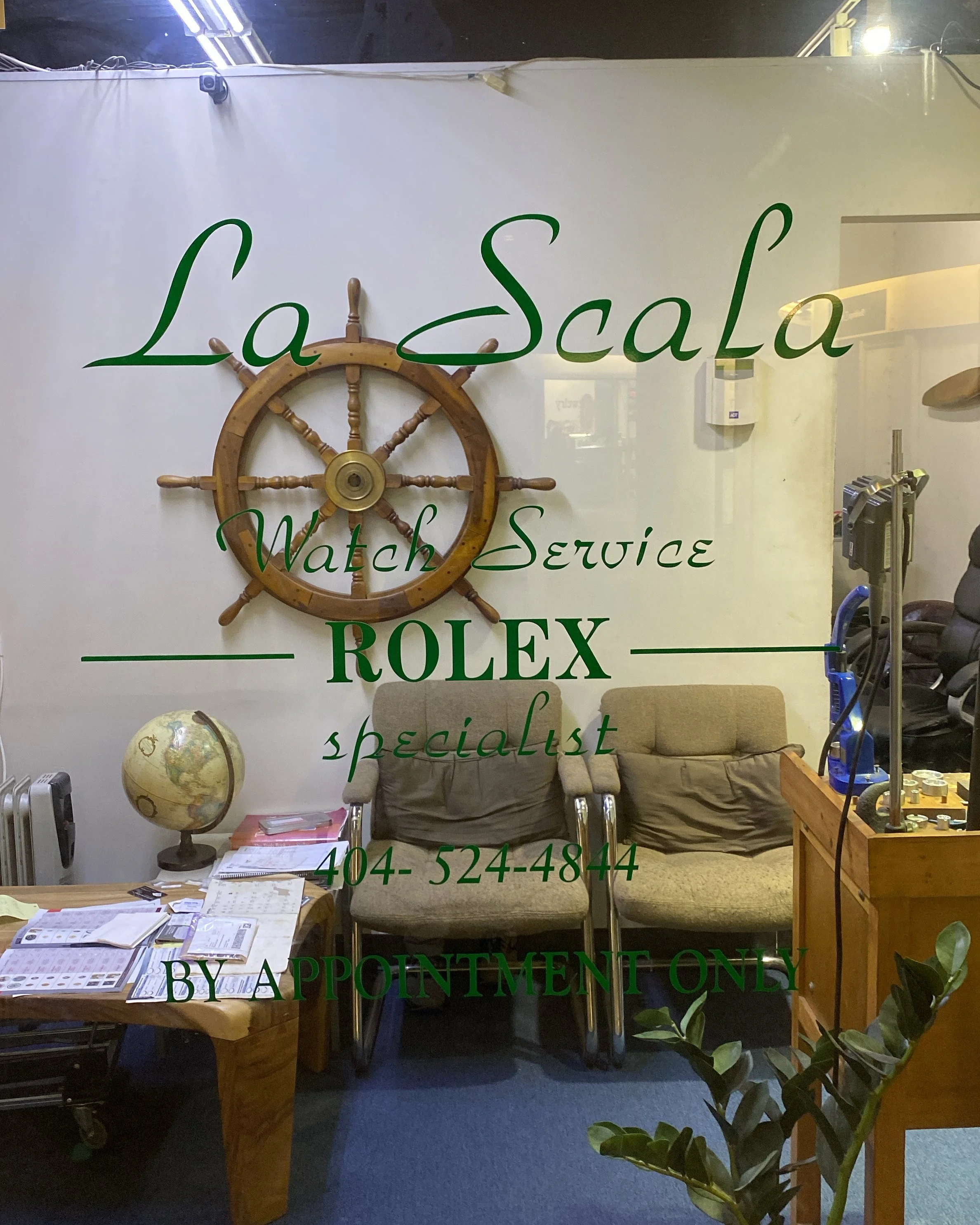 Store window with green lettering advertising watch servicing and Rolex, featuring a wooden ship's wheel, a globe, a desk with papers, a beige couch, and interior furnishings.