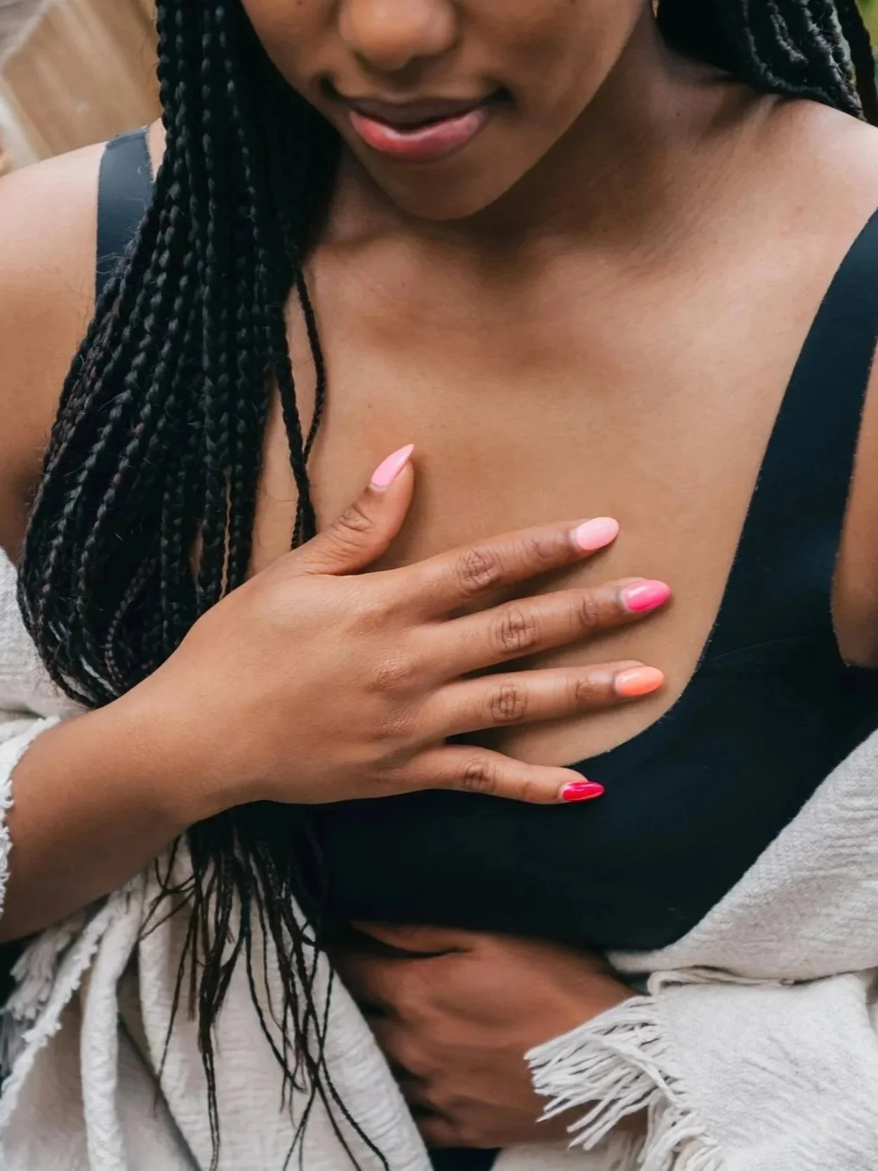 Close-up of a woman with braids touching her chest with her hand, which has multicolored pink and red nail polish, while wearing a black top and a light-colored fringed shawl.