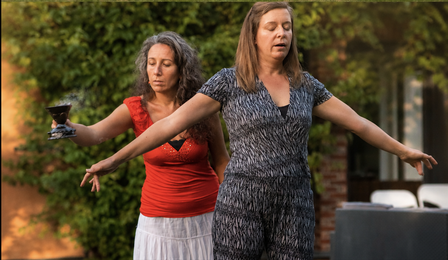 Two women practicing outdoor yoga, with one woman in a red top and white skirt, and the other in a patterned jumpsuit, standing with arms extended and eyes closed.