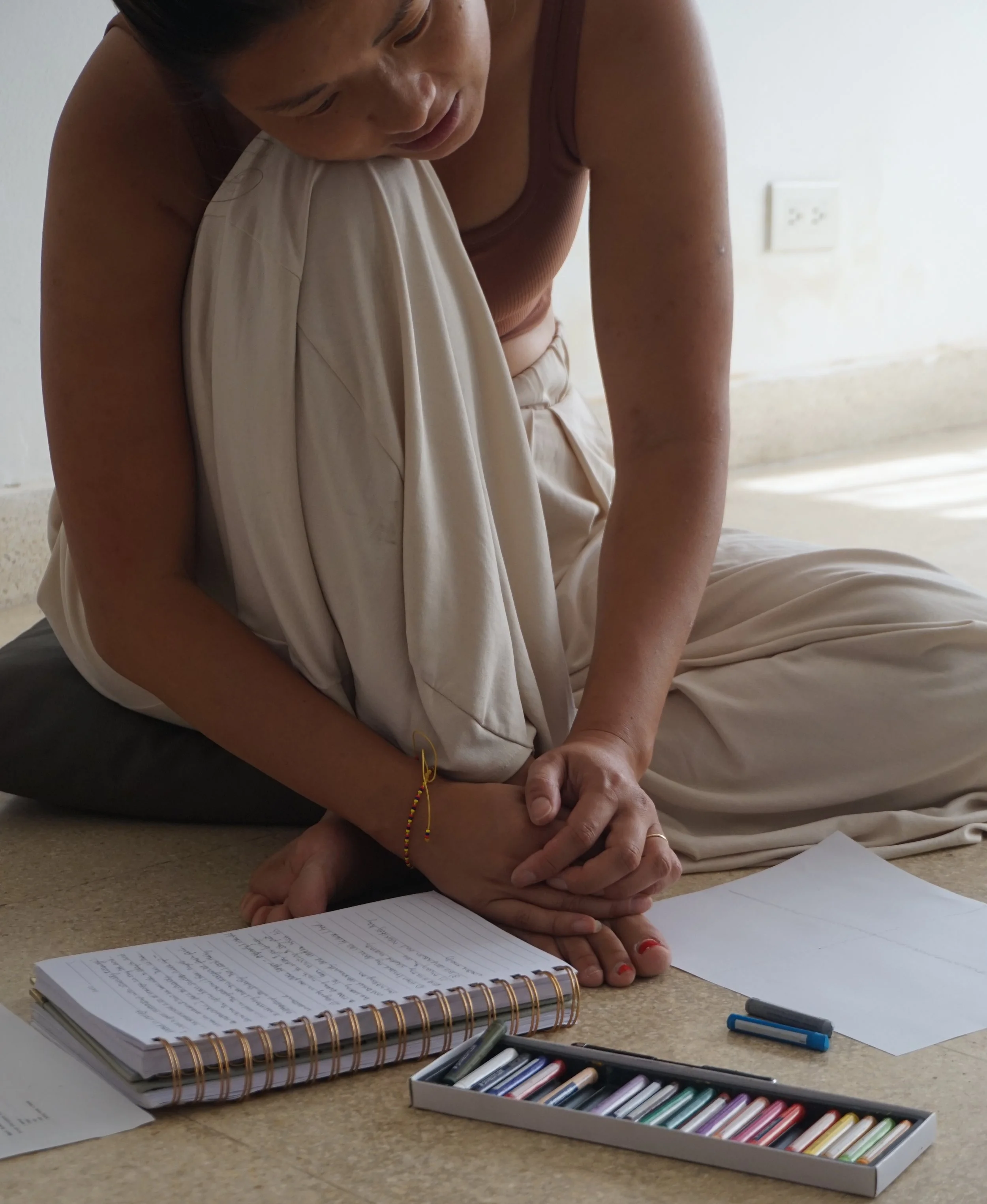 A woman sitting on the floor, leaning over a table, writing on a sheet of paper surrounded by notebooks, colored pastels, and marker pens.
