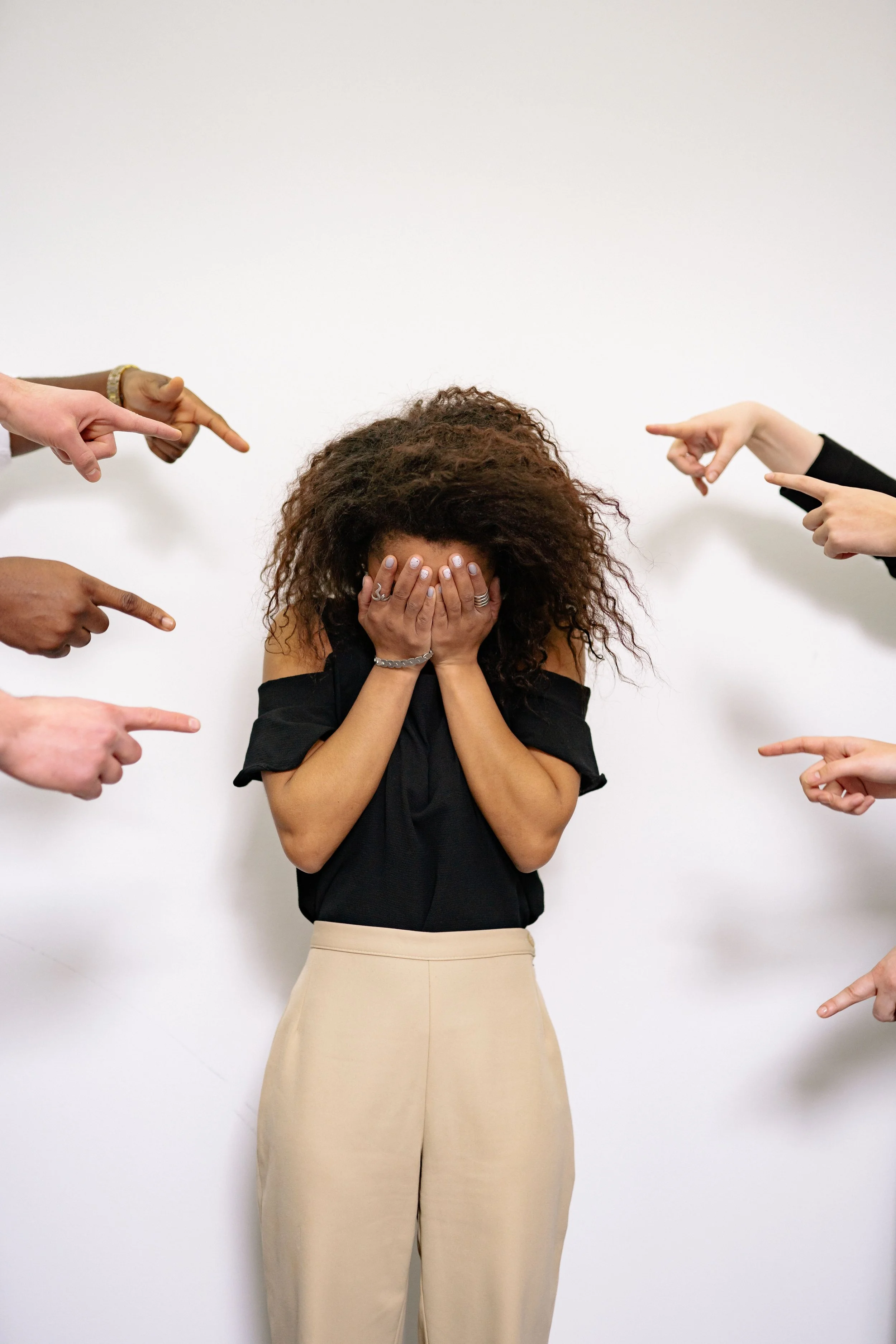 A woman with curly hair covering her face with her hands surrounded by multiple hands pointing at her.