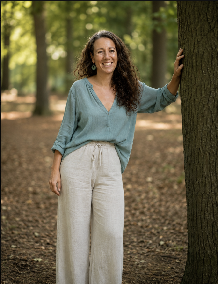 A woman with long curly brown hair smiling and leaning against a tree in a forest, wearing a light blue top and beige pants.