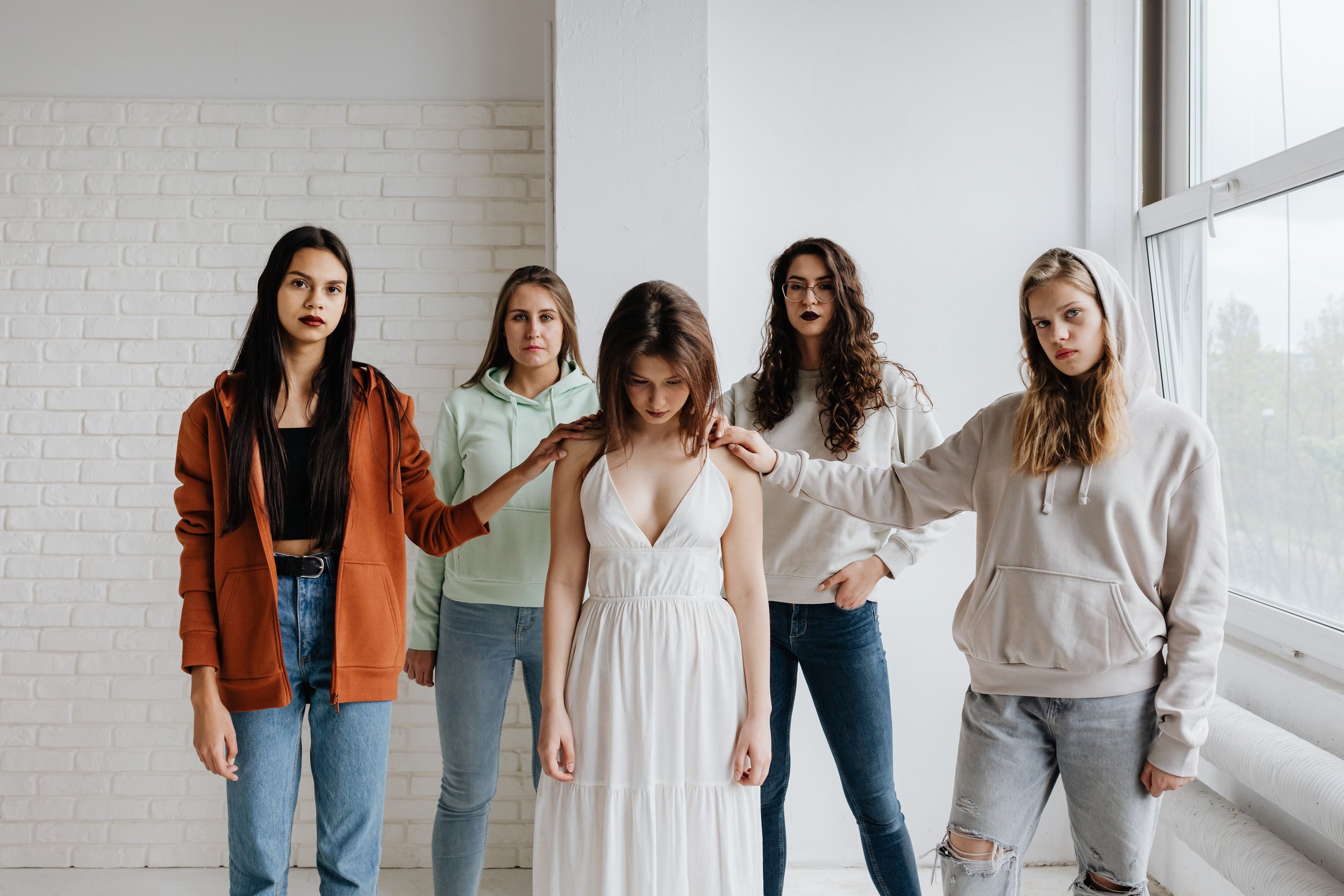 Five young women standing together in a bright room with white brick and plain walls, some with their hands on a girl in a white dress who is looking down.