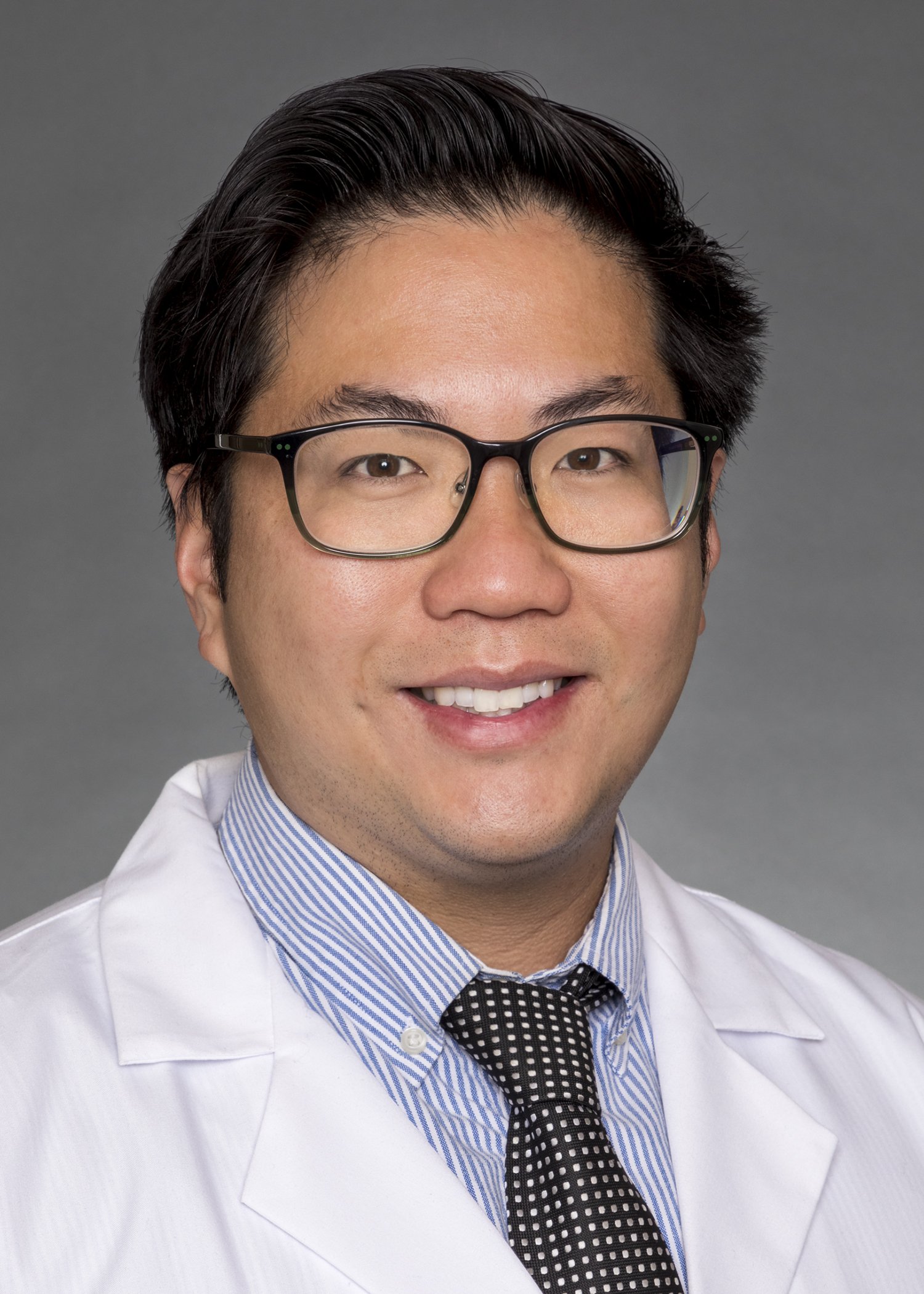 A male doctor of Asian descent smiling, wearing glasses, a white coat, a light blue striped dress shirt, and a black and white dotted tie, against a gray background.