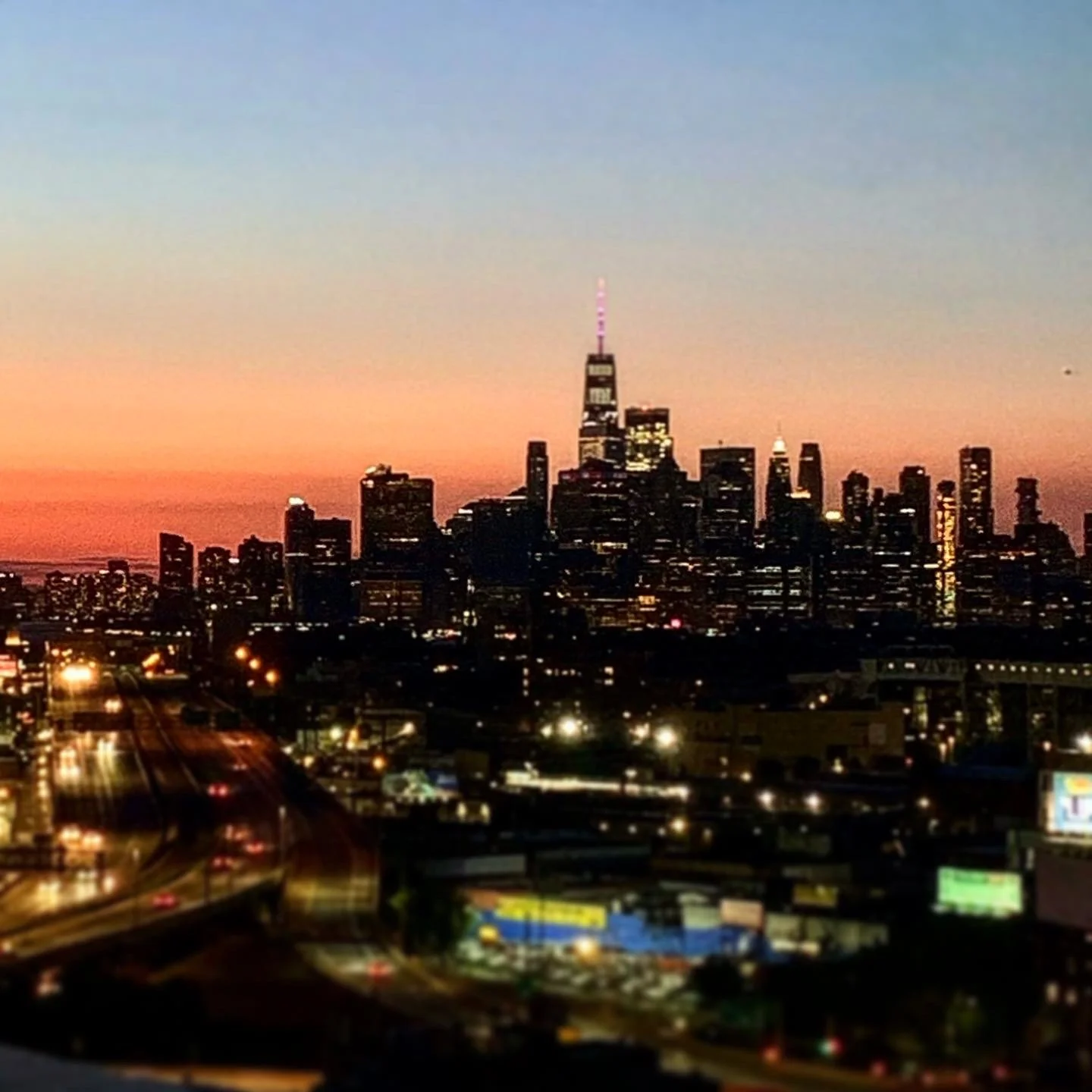 Skyline of a city during sunset with high-rise buildings, including a prominent tower with a lit antenna, and city lights beginning to turn on.