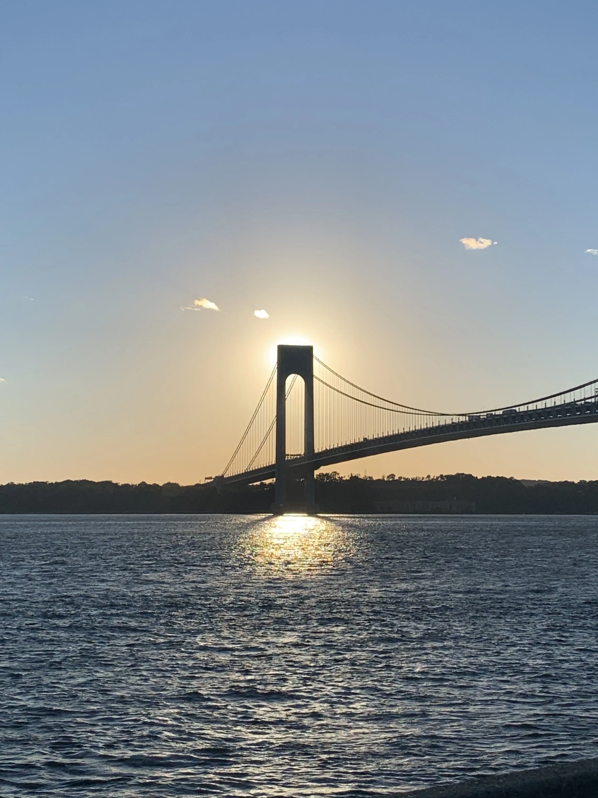 A suspension bridge with towers is seen at sunset over a body of water, with the sun's glow around the towers and a few scattered clouds in the sky.