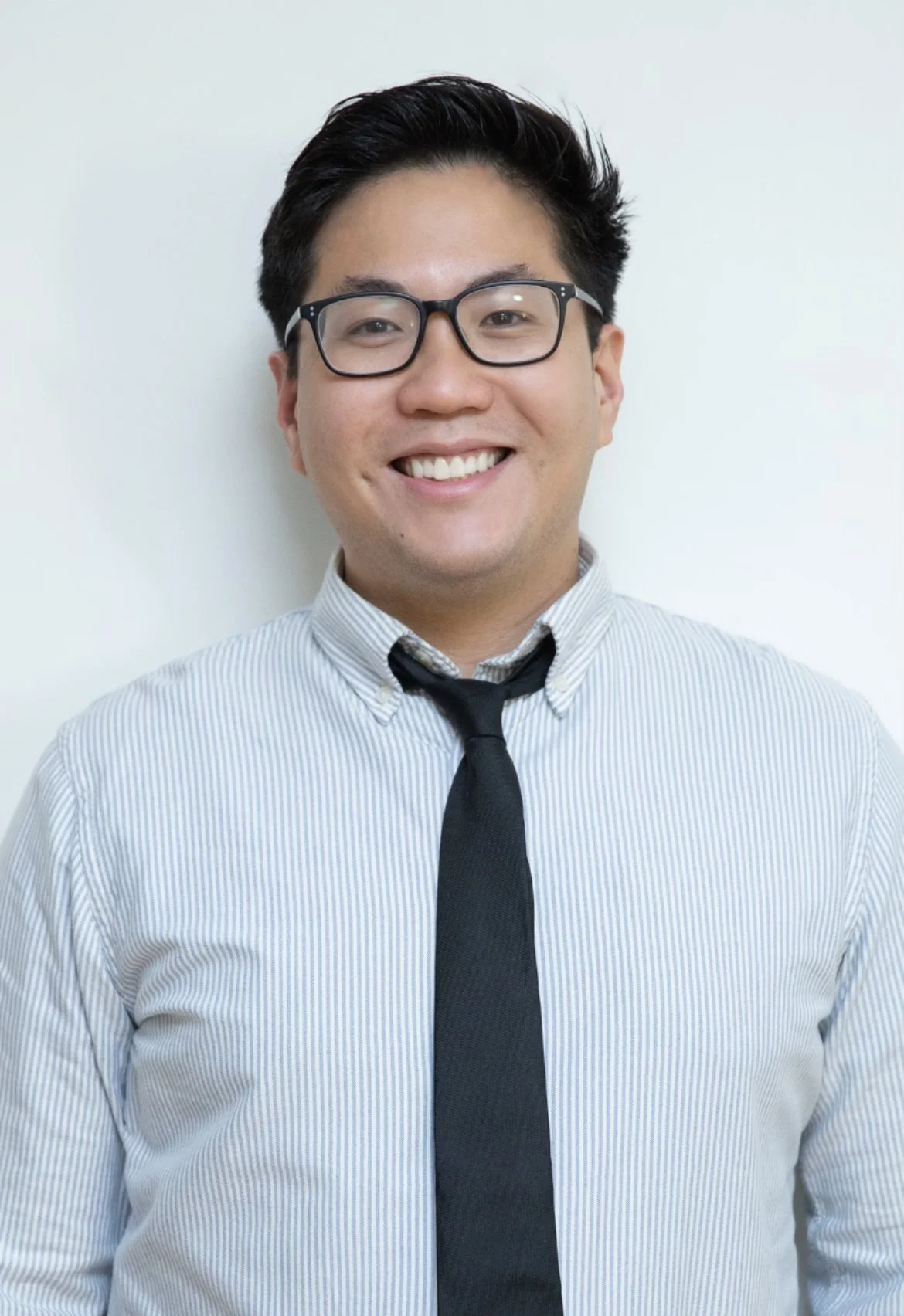 Smiling young man wearing glasses, a white striped shirt, and a black tie against a plain light-colored background.