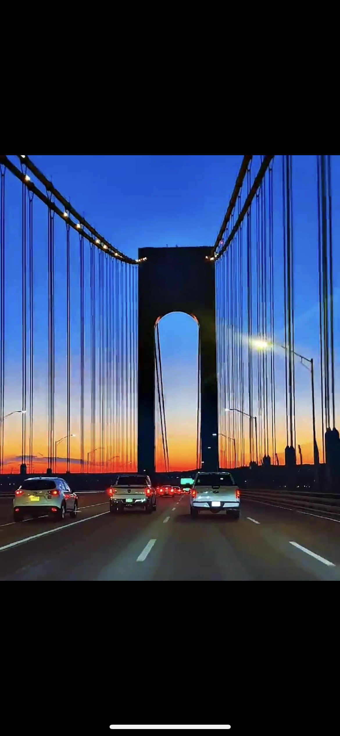 Cars driving on a bridge at sunset with a large dark tower structure in the background and a clear sky transitioning from orange to blue.