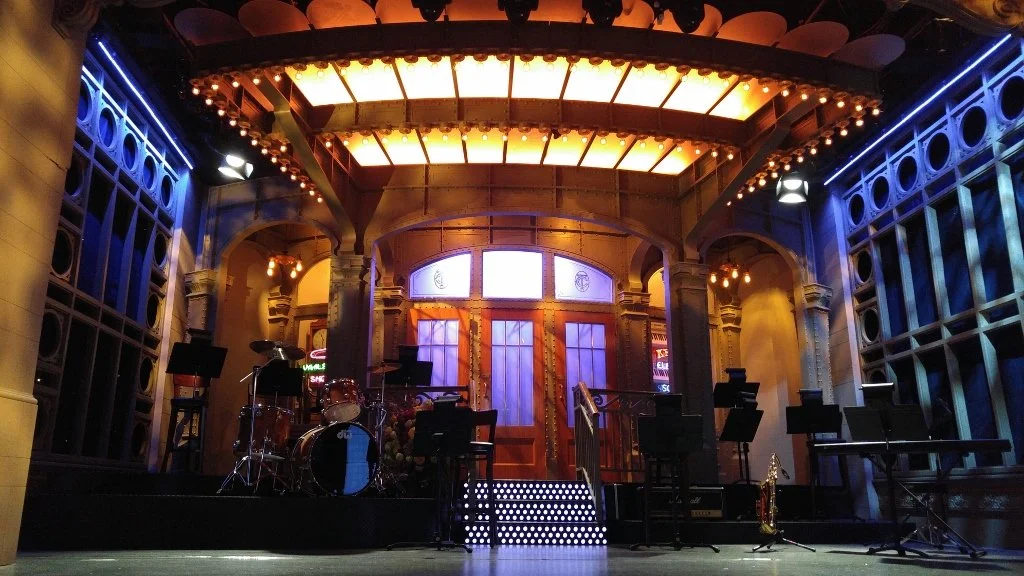 Empty stage with musical instruments, including drums, keyboards, and microphones, set against a decorated theater-style backdrop with warm lighting and neon signs.