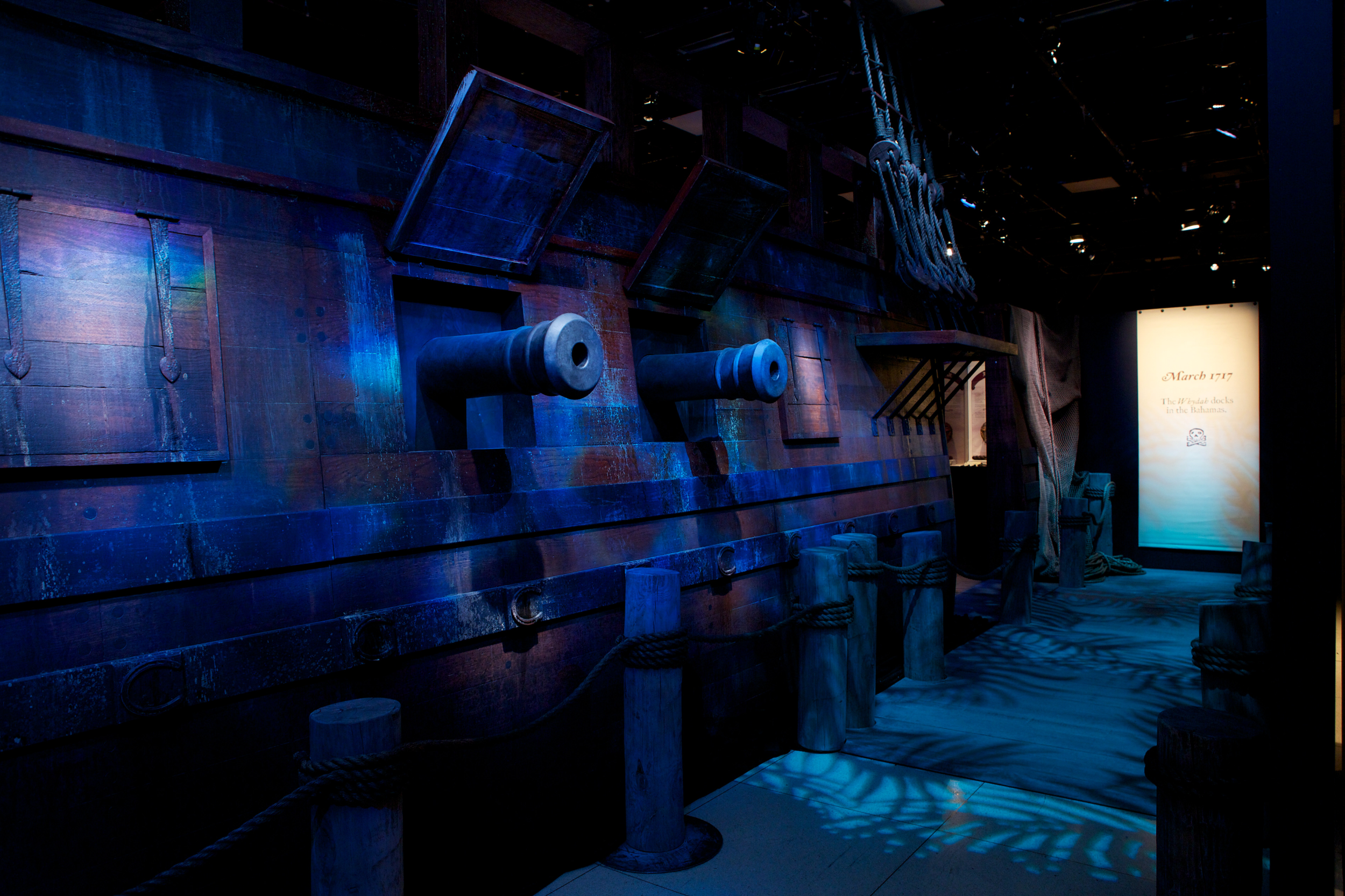 A display of a wooden pirate ship with cannons, ropes, and ladders, lit with blue lighting at a museum exhibit.