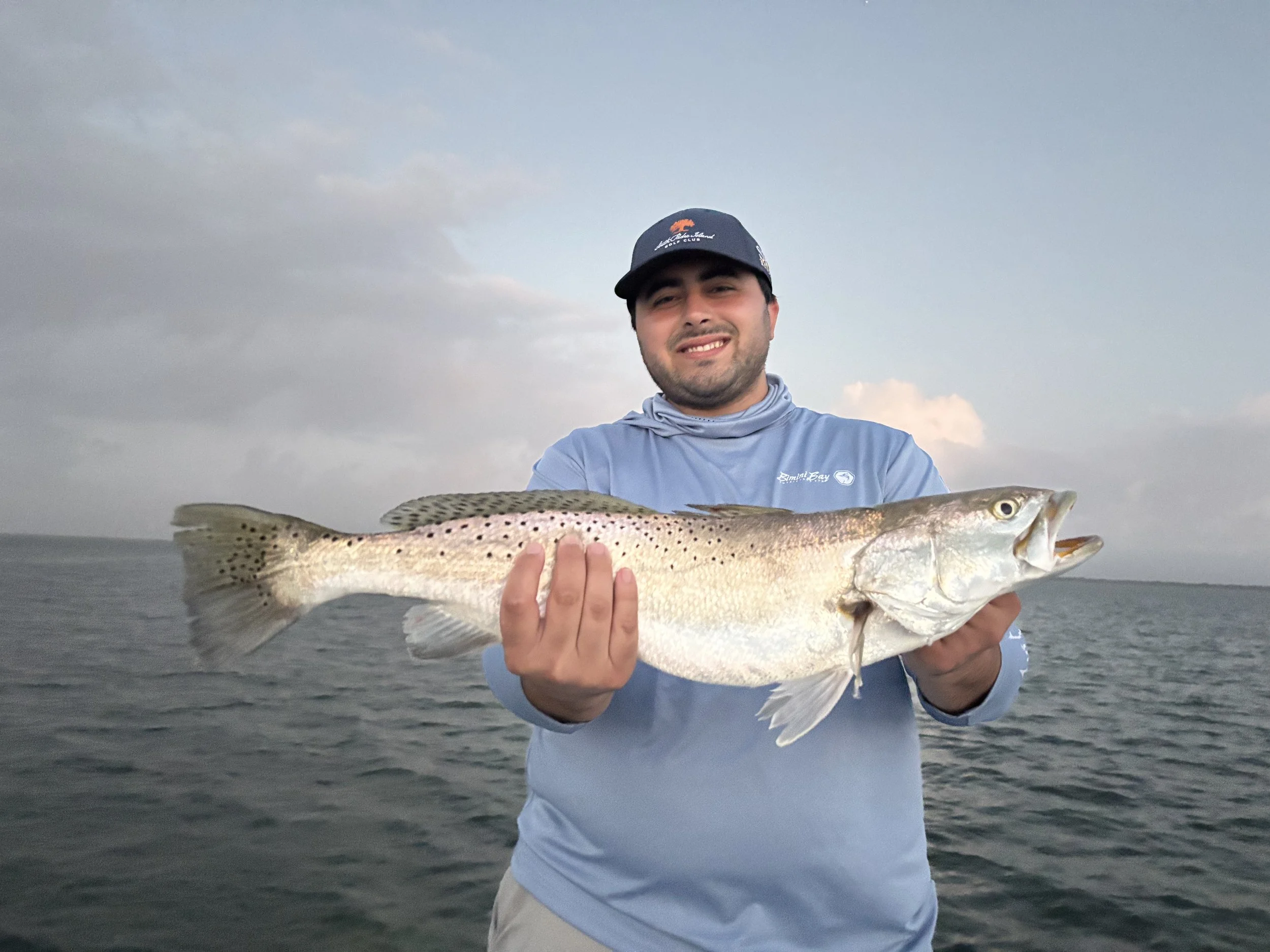 A young man wearing a blue long sleeve shirt and a cap holding a large fish over water during sunset
