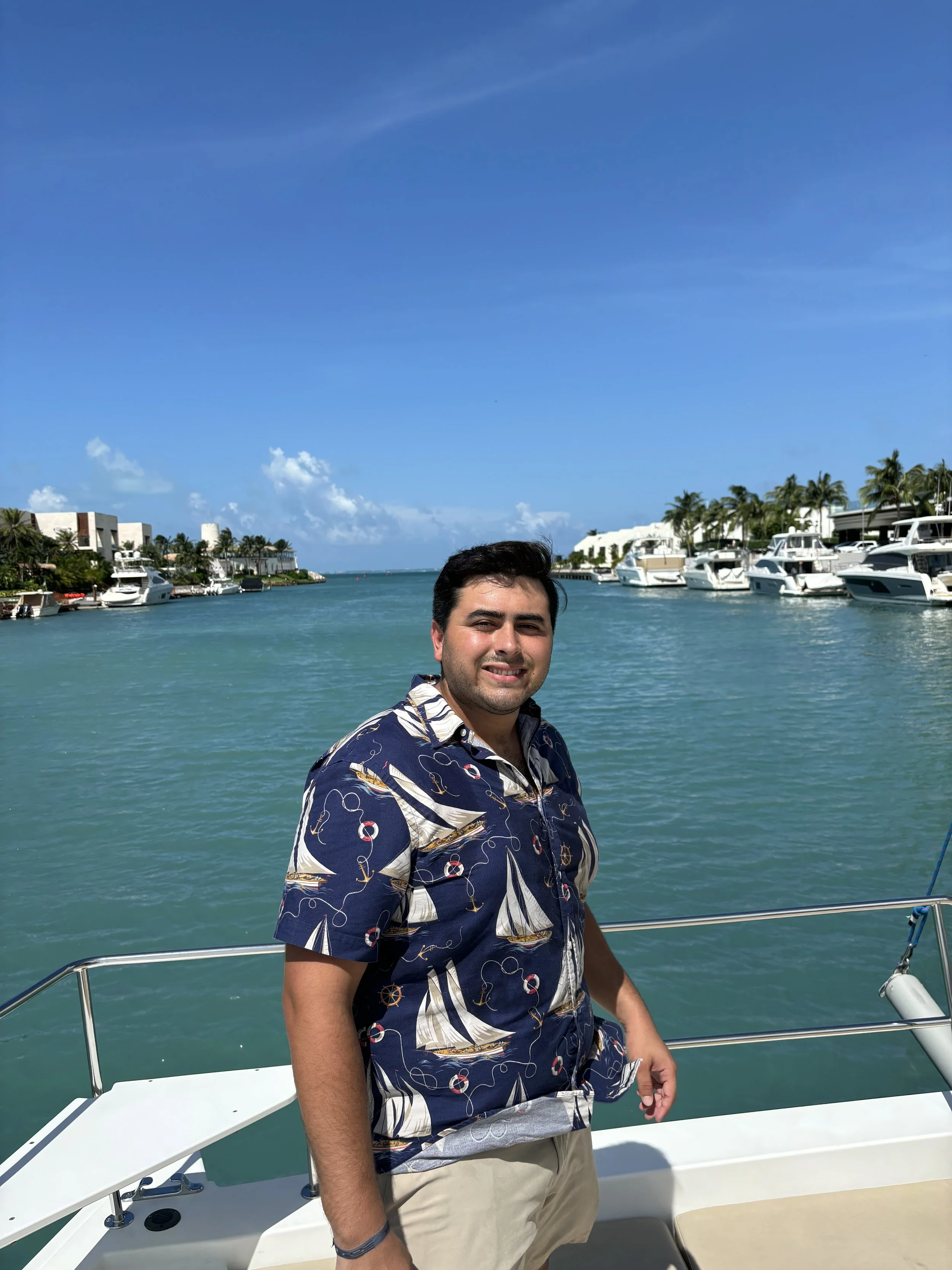 A man smiling on a boat with a marina and yachts in the background under a clear blue sky