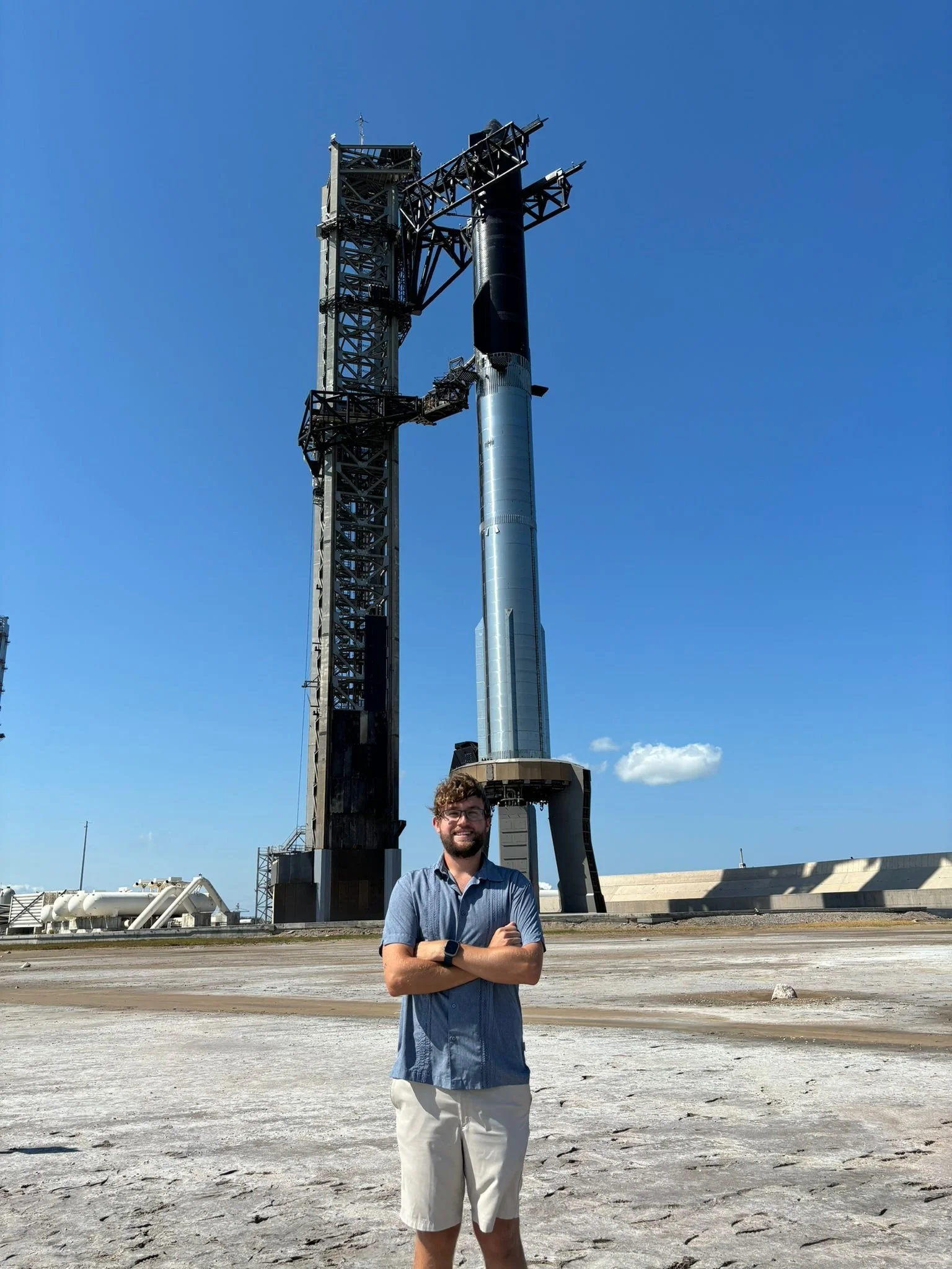 A man with curly hair and glasses standing outdoors with arms crossed in front of a spaceX rocket launching pad, under a clear blue sky.