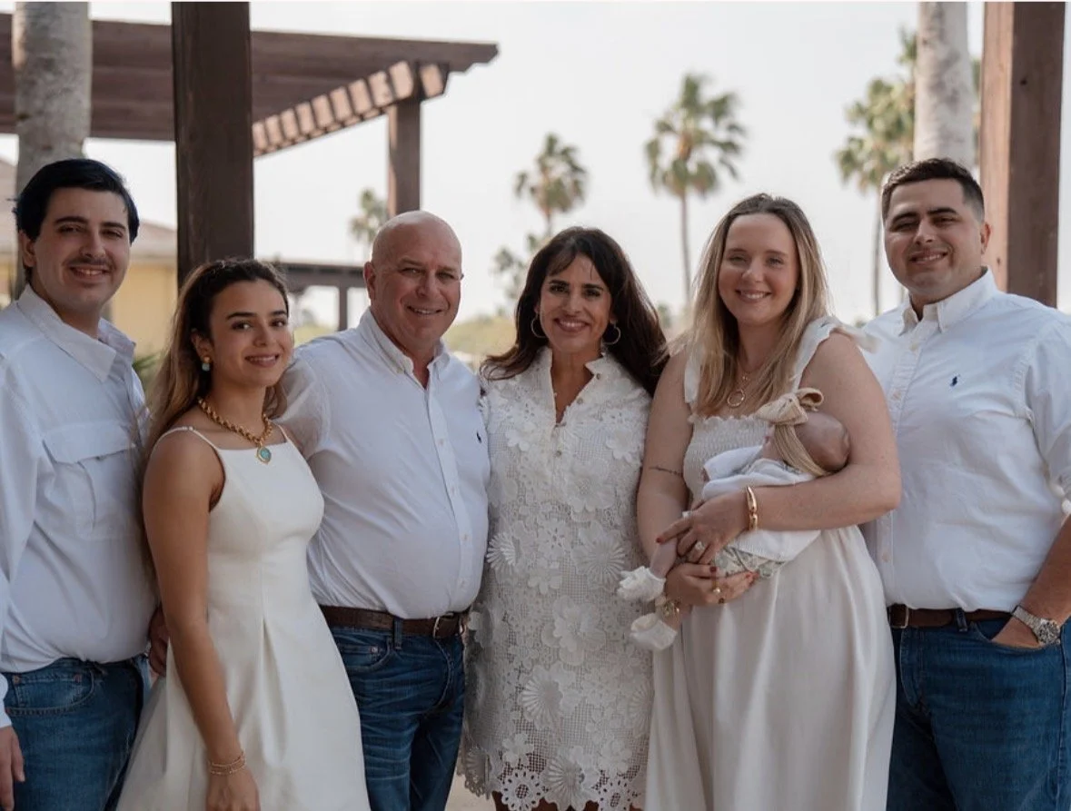A group of seven people, including a baby, posing outdoors under a wooden pergola with palm trees in the background. Everyone is dressed in white or light-colored clothing and smiling at the camera.