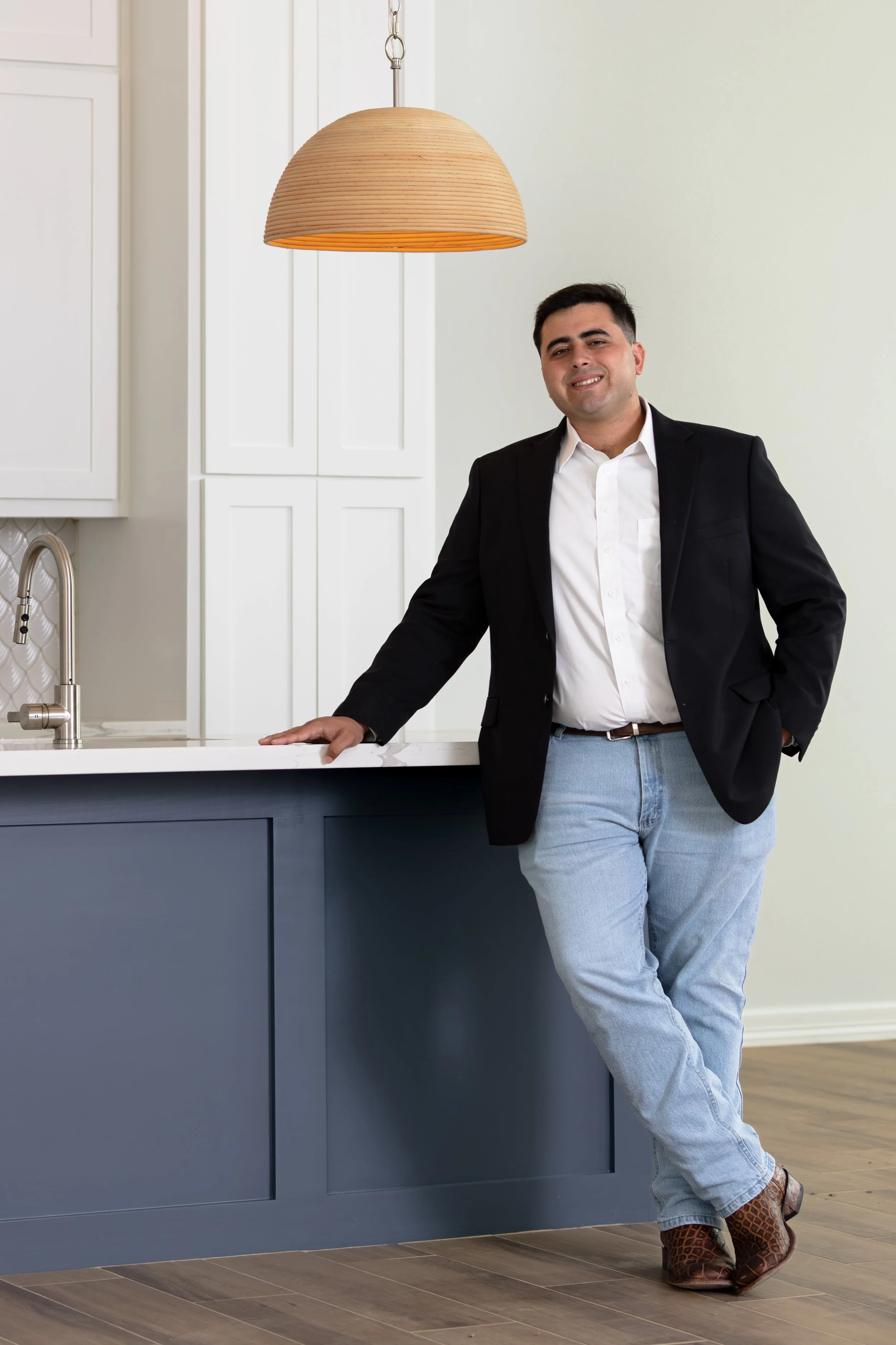 Man in black blazer and light blue jeans standing in a modern kitchen with dark blue cabinets and white walls, leaning on the counter and smiling.
