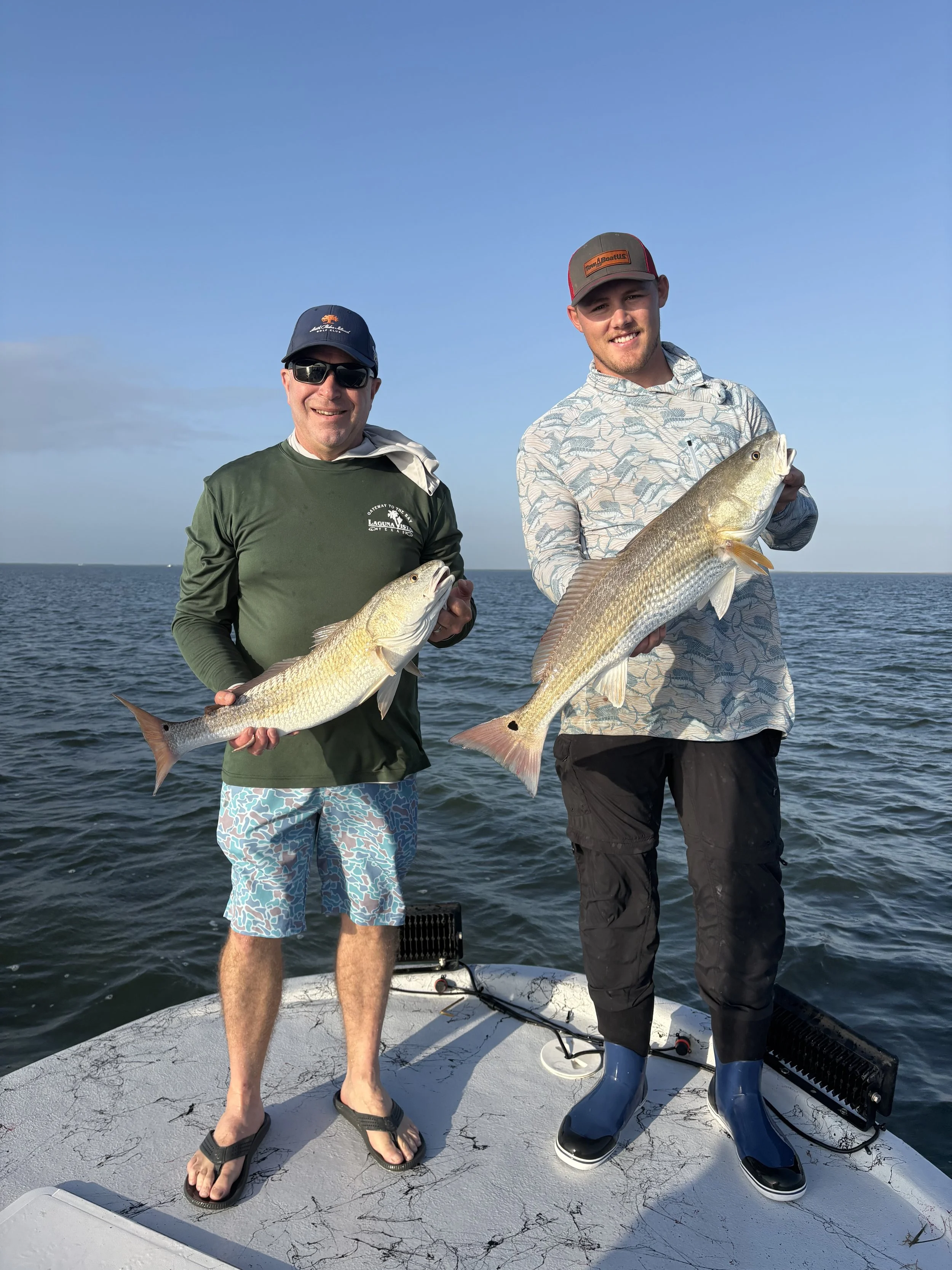 Two men standing on a boat holding large fish with an ocean and clear sky in the background.