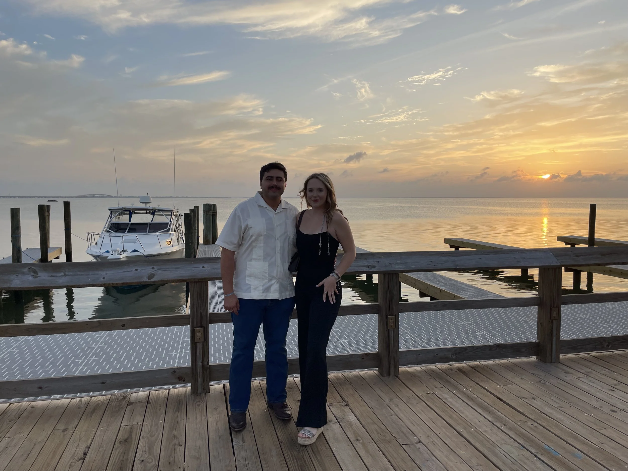 A couple stands on a wooden dock at sunset by the water, with a boat and poles in the background.