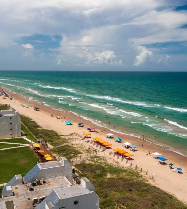 Aerial view of a beach with colorful umbrellas, lounge chairs, and people near the shoreline under partly cloudy skies.
