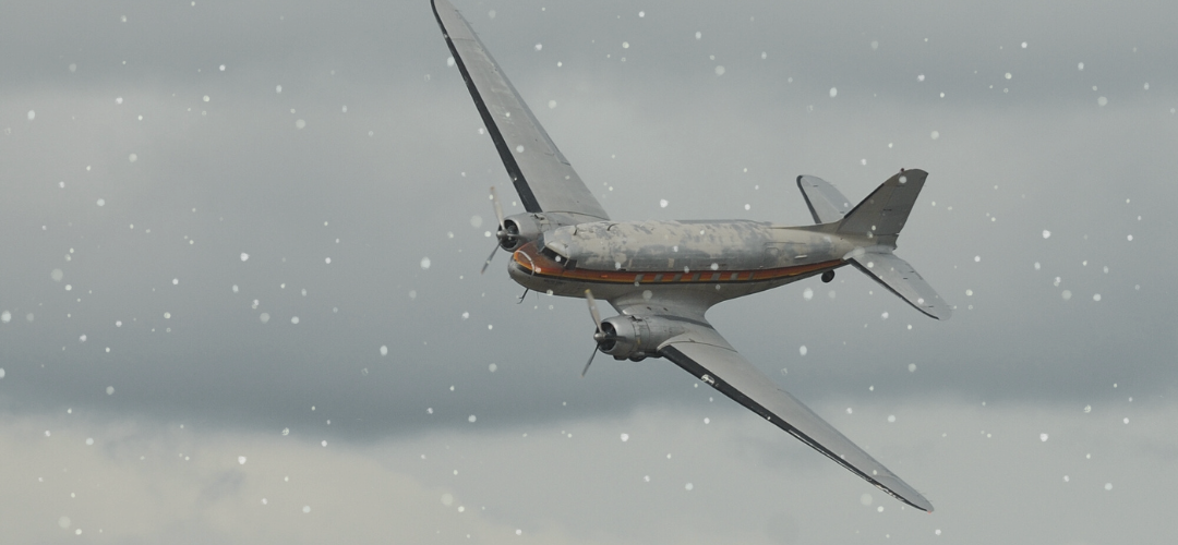 A vintage twin-engine propeller airplane flying through cloudy skies with snow falling.