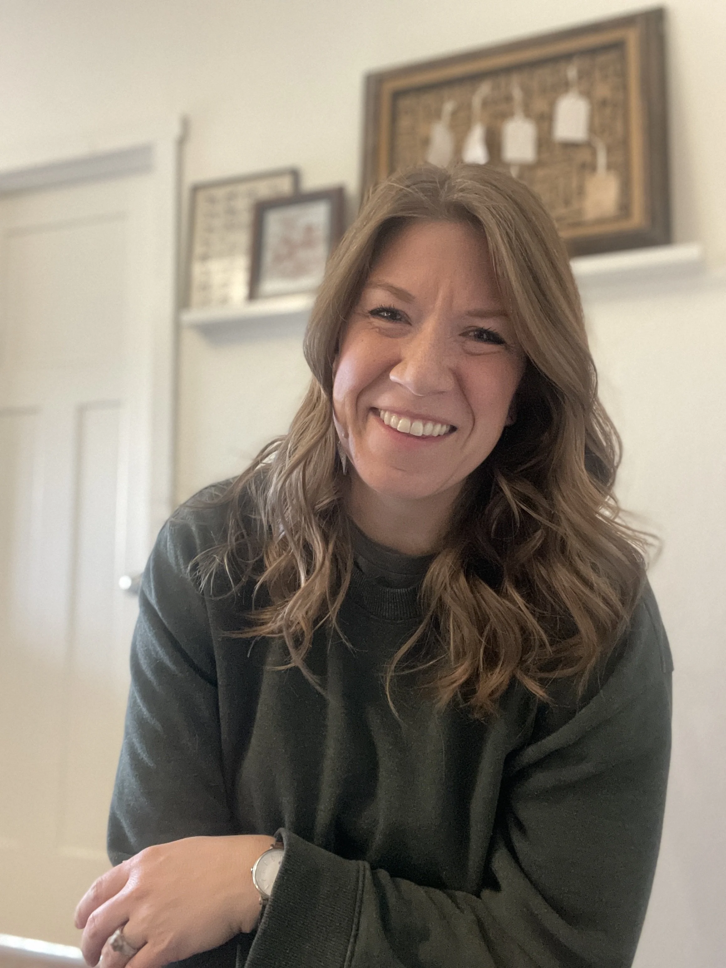 A woman with long wavy copper hair smiling, wearing a green sweater and a watch, sitting indoors with a blurry background that includes framed artwork on a wall.