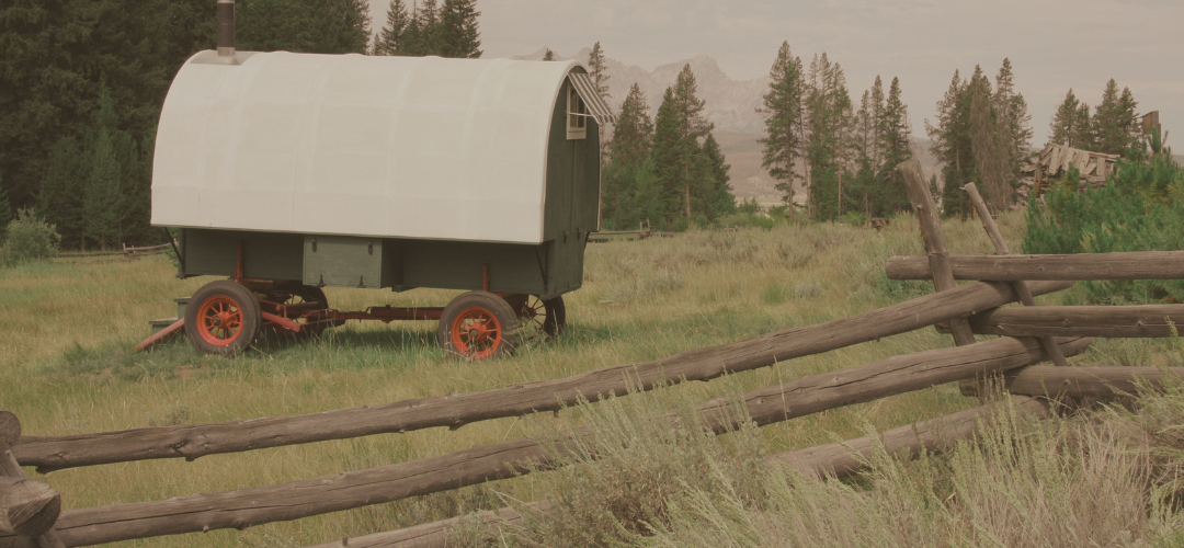A covered wagon with red wheels parked on a grassy field near a rustic wooden fence, with a forested area and mountains in the background.