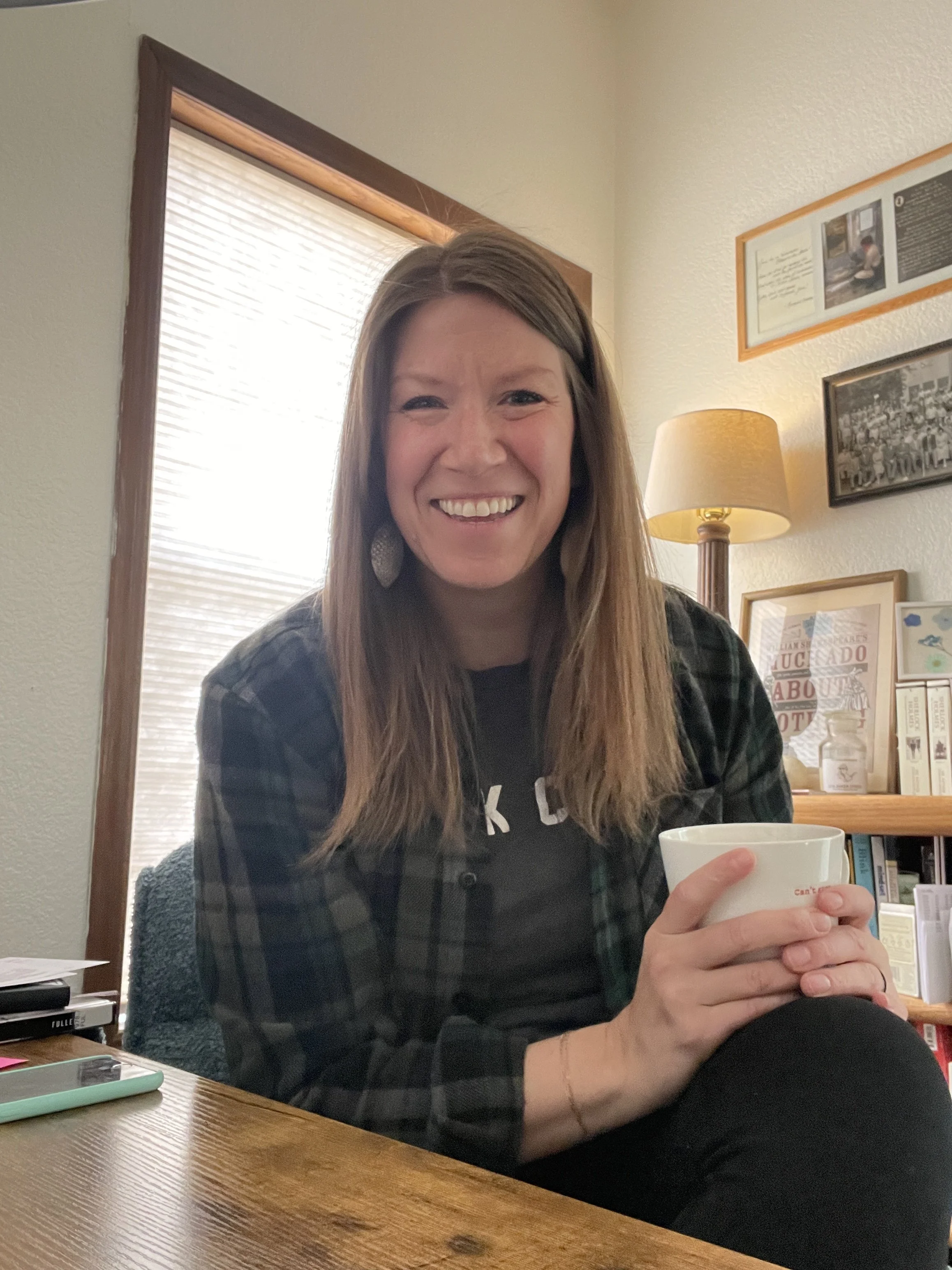 A woman with long copper hair smiling while holding a white mug, sitting at a wooden table in a cozy, well-lit room with framed pictures and books on the wall.