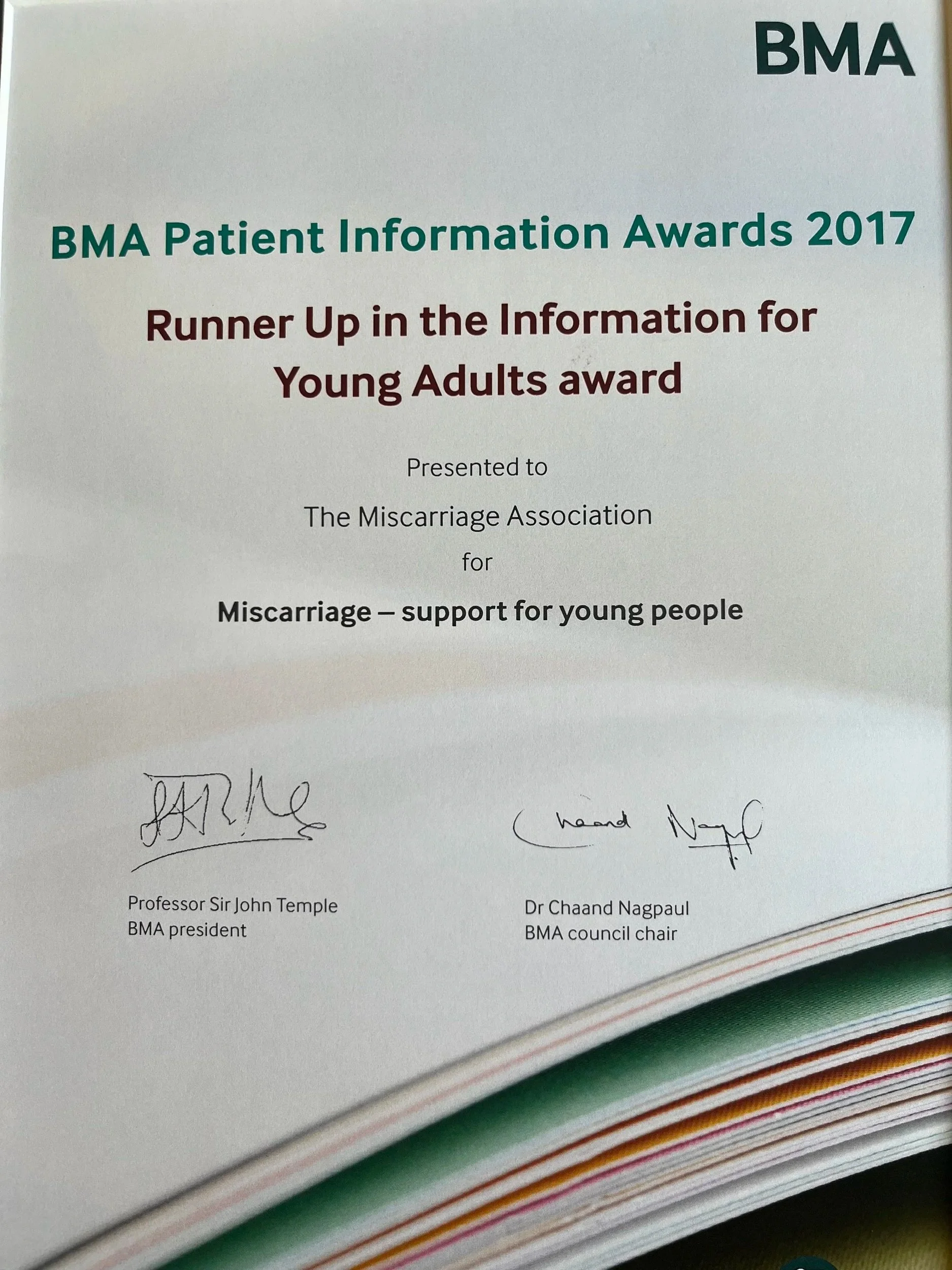 Close-up photo of a certificate titled 'BMA Patient Information Awards 2017' with the subtitle 'Runner Up in the Information for Young Adults award'. The certificate is presented to The Miscarriage Association for miscarriage support for young people. It is signed by Professor Sir John Temple and Dr. Chaand Nagpal and is placed on top of a stack of colorful papers.