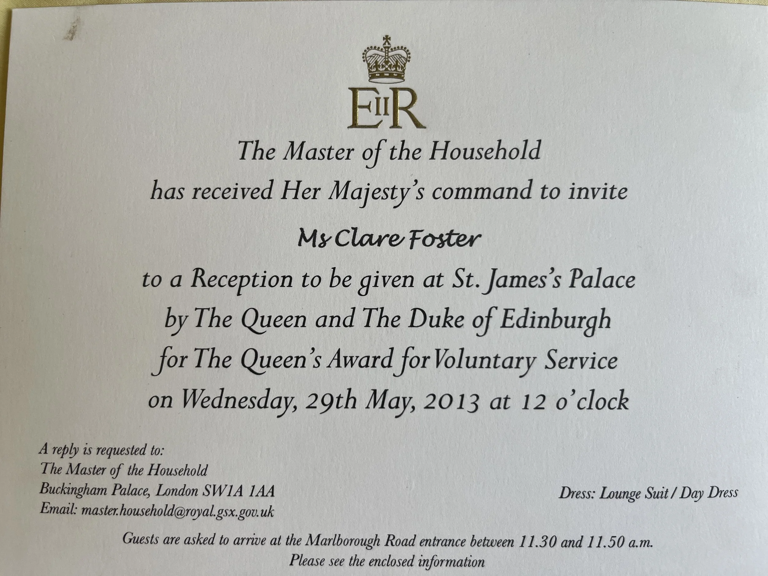 Invitation card with a royal crown graphic at the top, reading 'The Master of the Household has received Her Majesty's command to invite Ms. Clare Foster to a Reception at St. James's Palace on Wednesday, 29th May 2013 at 12 o'clock. Dress code: Lounge Suit / Day Dress.'
