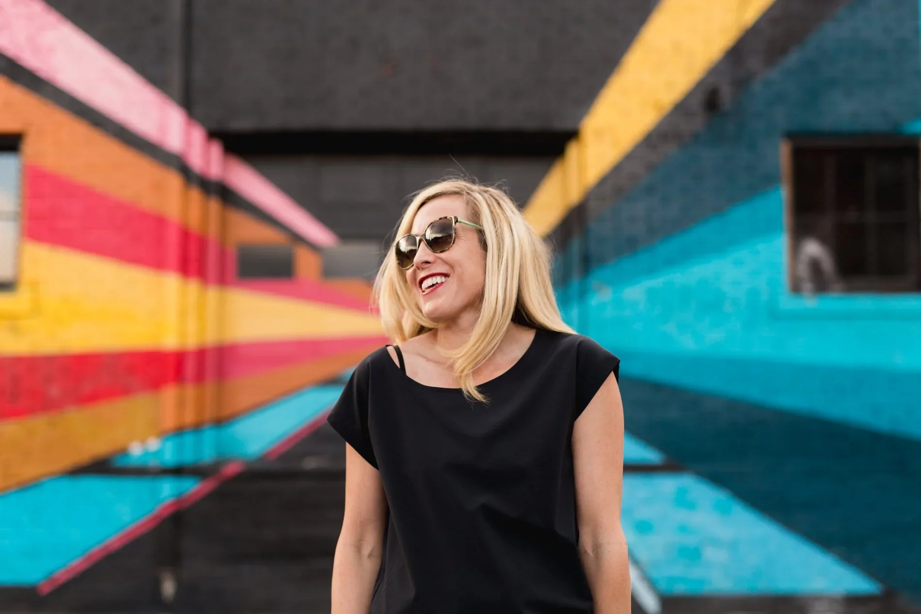 A woman with blonde hair wearing large sunglasses and a black top, smiling and standing in front of a colorful mural with diagonal blue, yellow, pink, peach, and red stripes.
