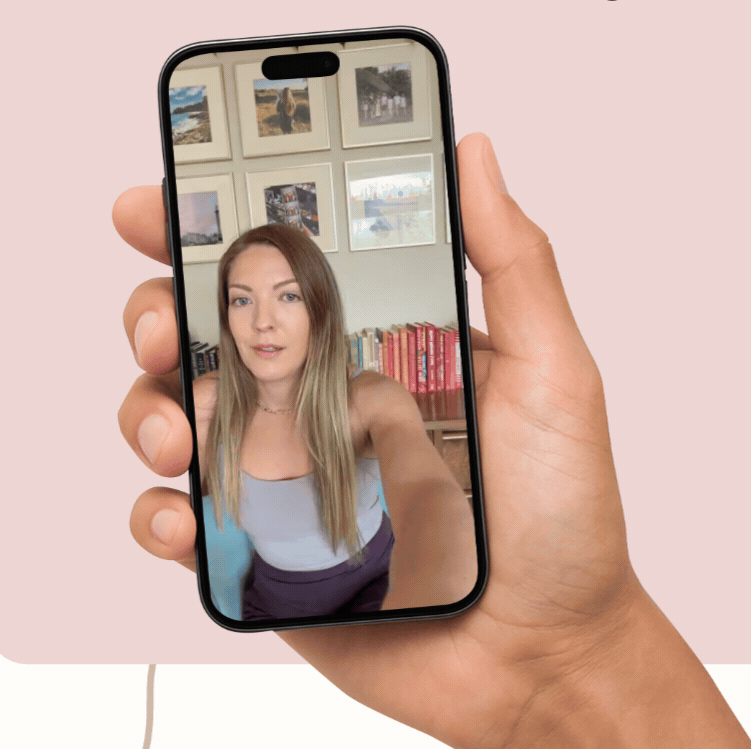 A person holding a smartphone capturing a selfie of a woman with long blonde hair, indoors with framed pictures on a wall and a bookshelf in the background.
