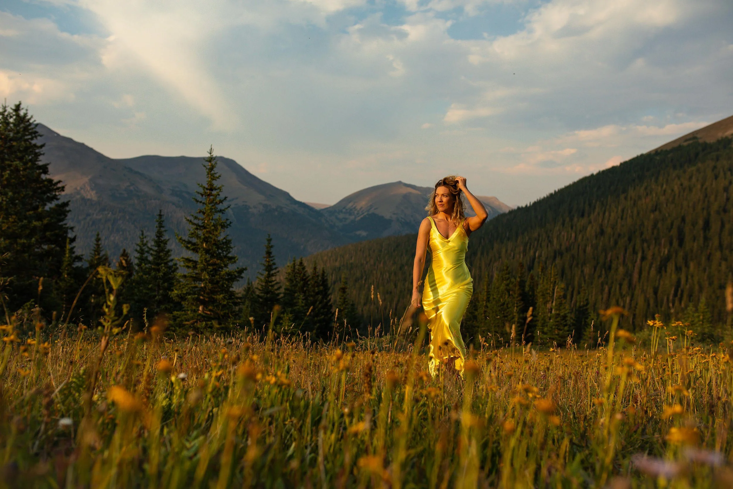 A woman in a yellow dress walking through a grassy field with yellow flowers, surrounded by tall pine trees and mountains in the background under a cloudy sky.
