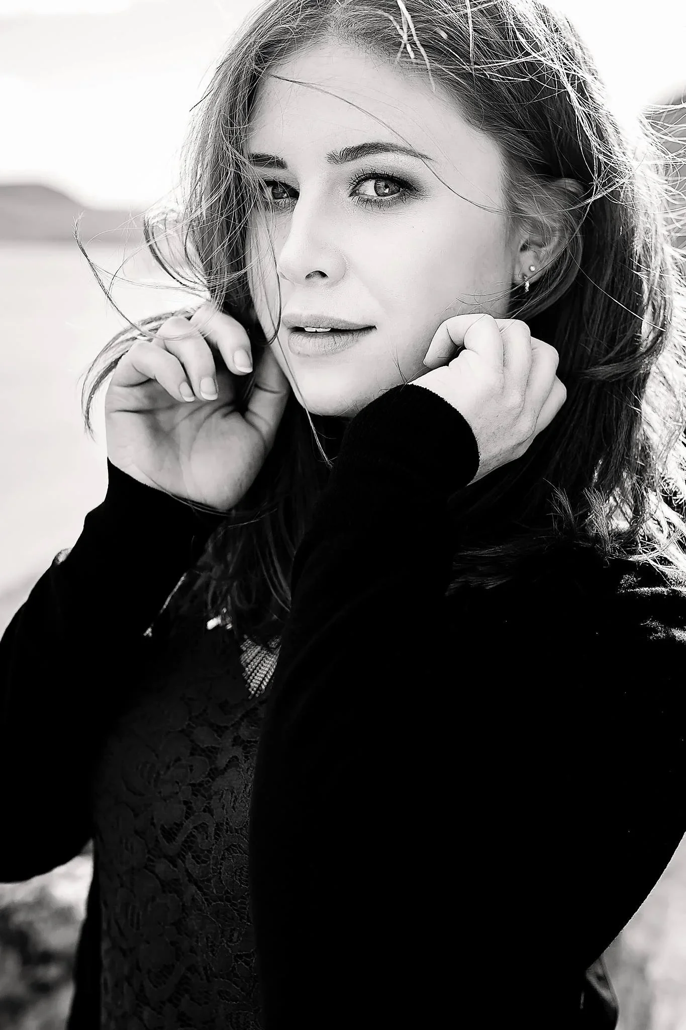 A black and white photo of a young woman with long hair, looking directly at the camera with one hand near her face, outdoors in natural light.