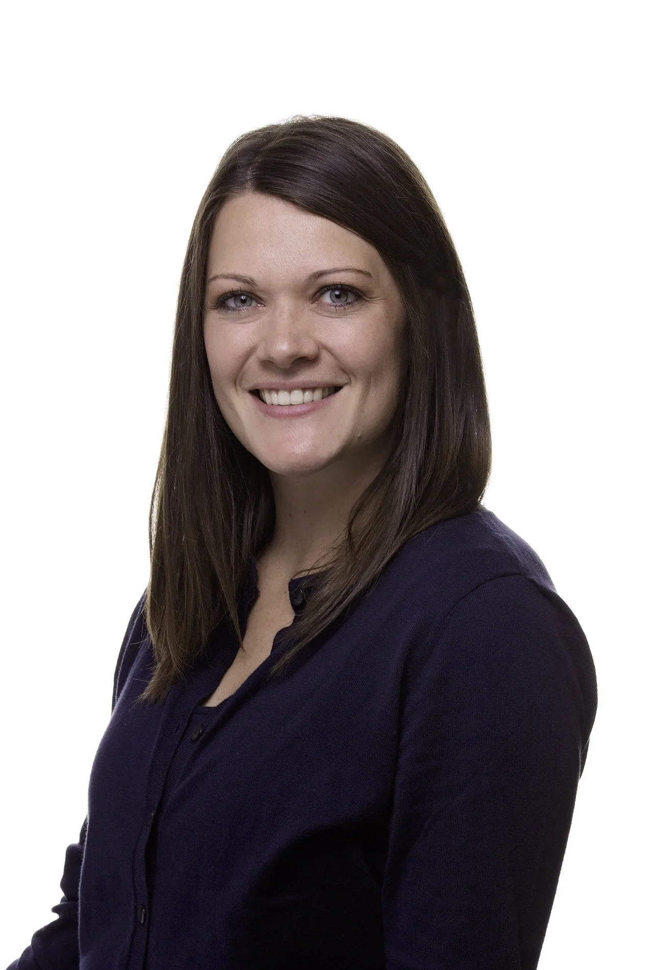 A woman with straight brown hair, blue eyes, smiling, wearing a dark navy blue blouse against a white background.