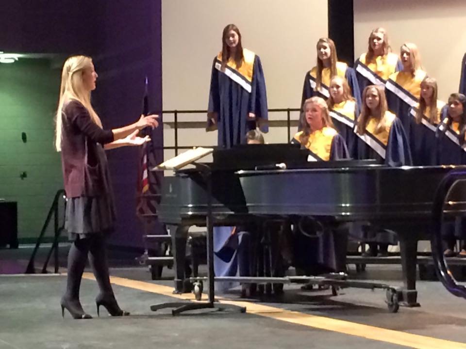 A woman conductor leading a children's choir performing on stage, with a grand piano in front of the choir.