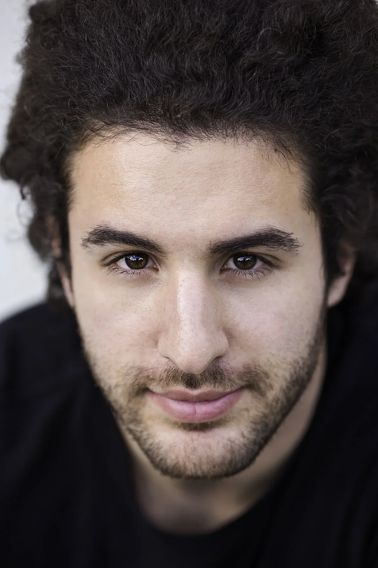 Close-up portrait of a man with curly brown hair, brown eyes, light beard, and mustache, wearing a black shirt.