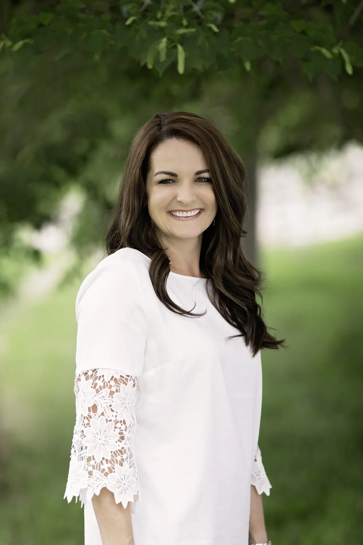 A woman with long dark hair smiling outdoors, wearing a white dress with lace sleeves, standing in front of green trees.