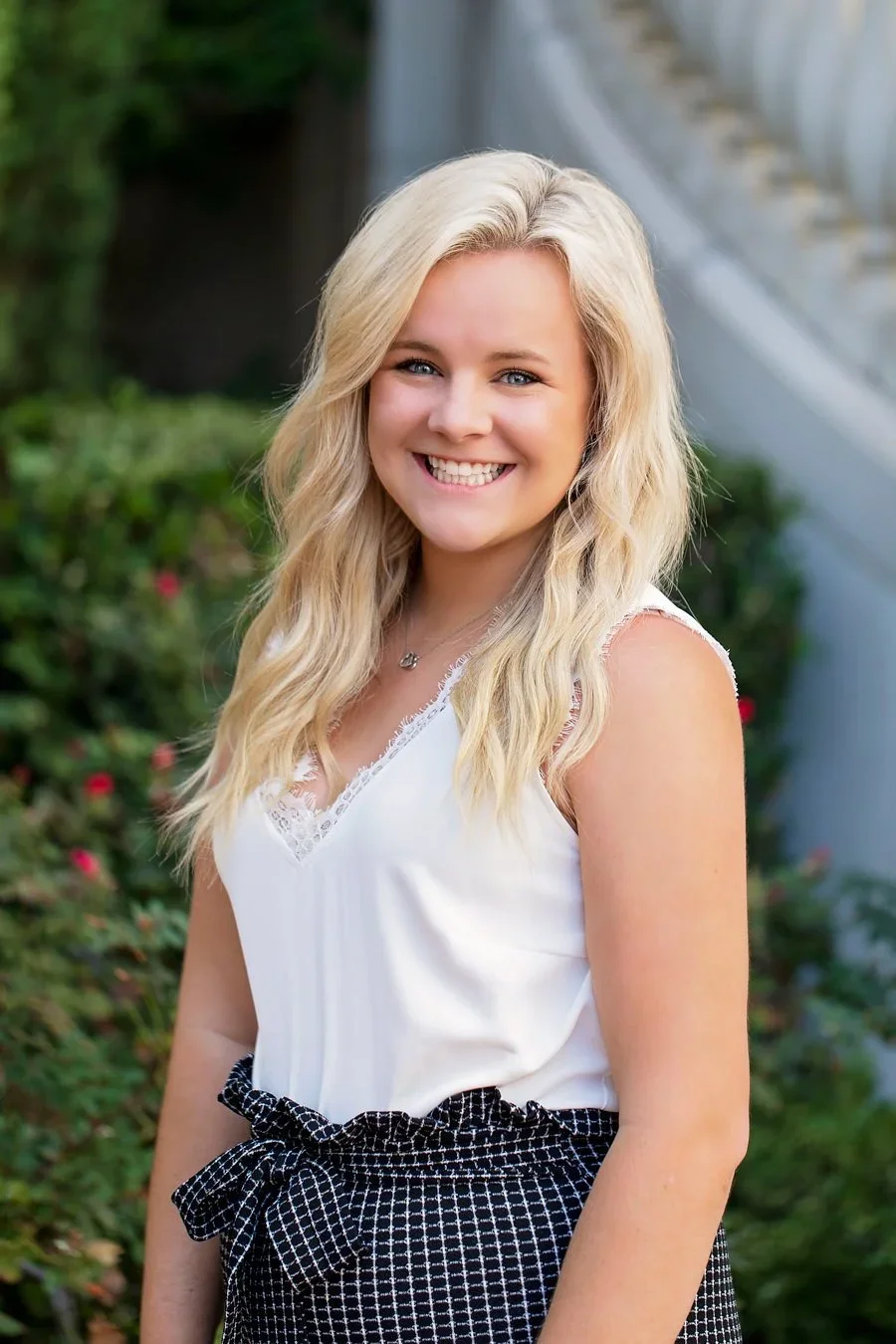A young woman with blonde hair smiling outdoors, wearing a white sleeveless top and black patterned skirt.