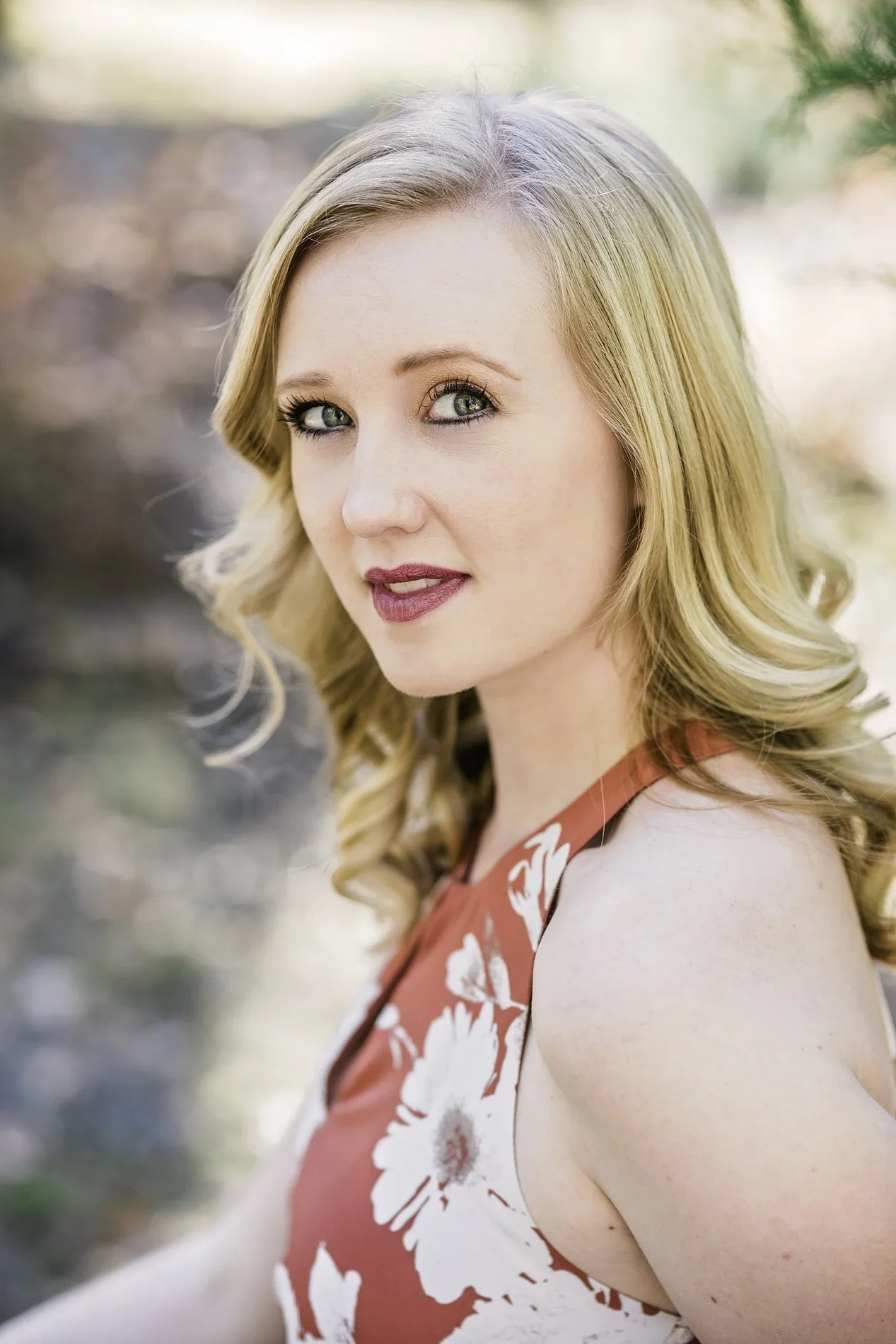 A woman with blonde hair and blue eyes wearing a red and white floral dress, outdoors with a blurred natural background.