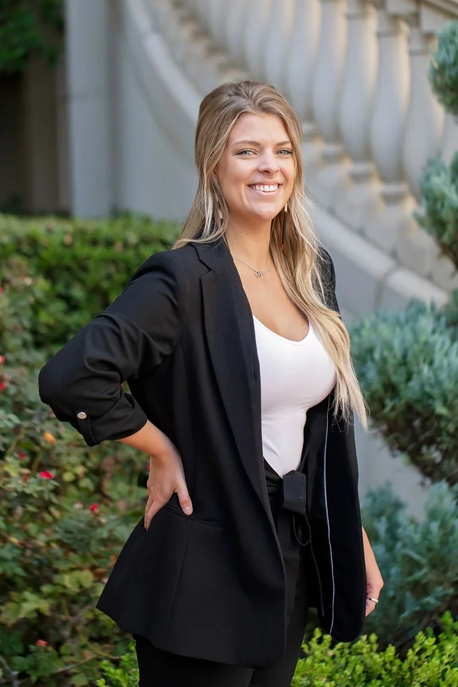 A smiling woman with long blonde hair, wearing a black blazer over a white top, standing outdoors near bushy plants and a stone staircase.