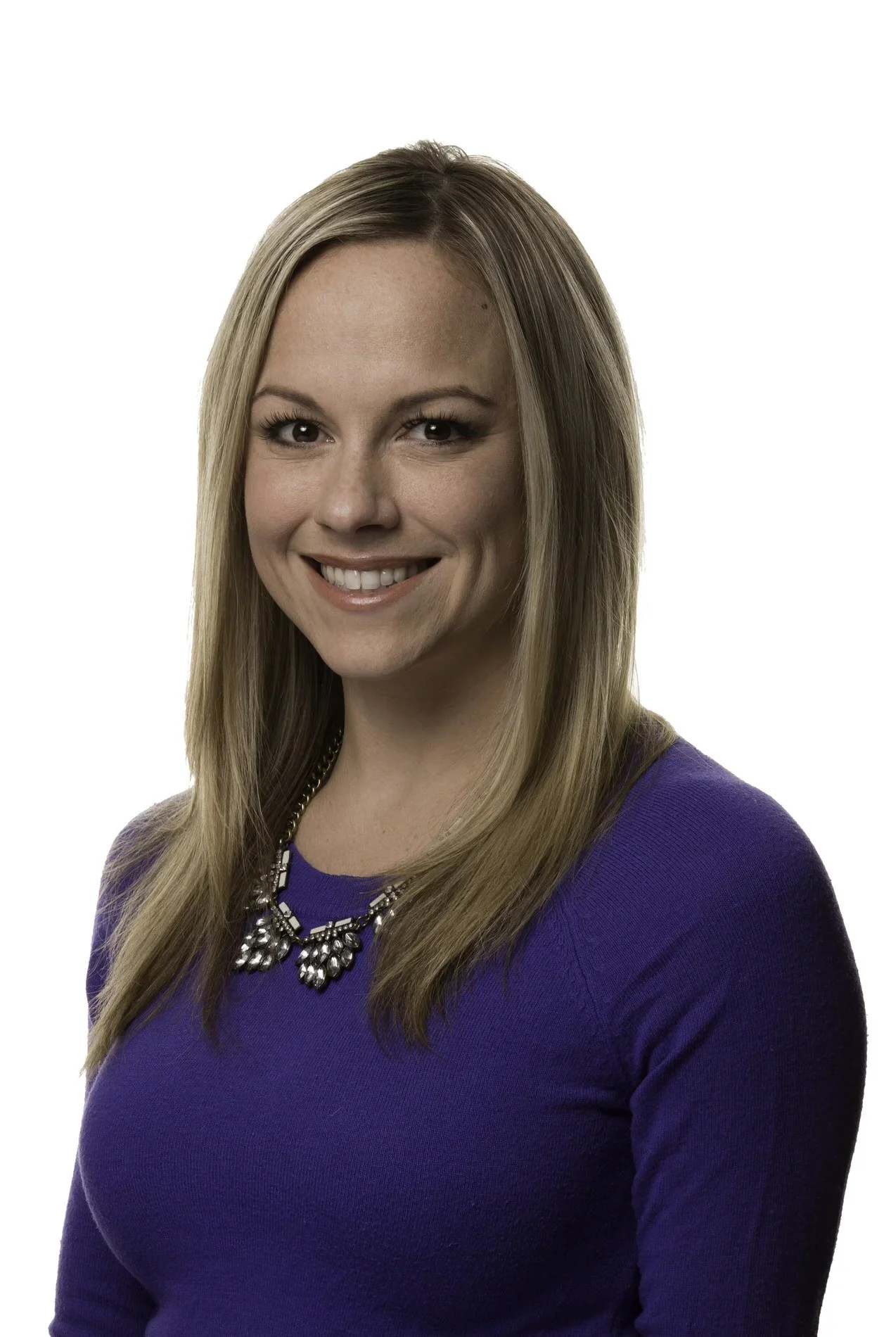 A woman with long blonde hair wearing a blue top and a statement necklace, smiling at the camera against a white background.