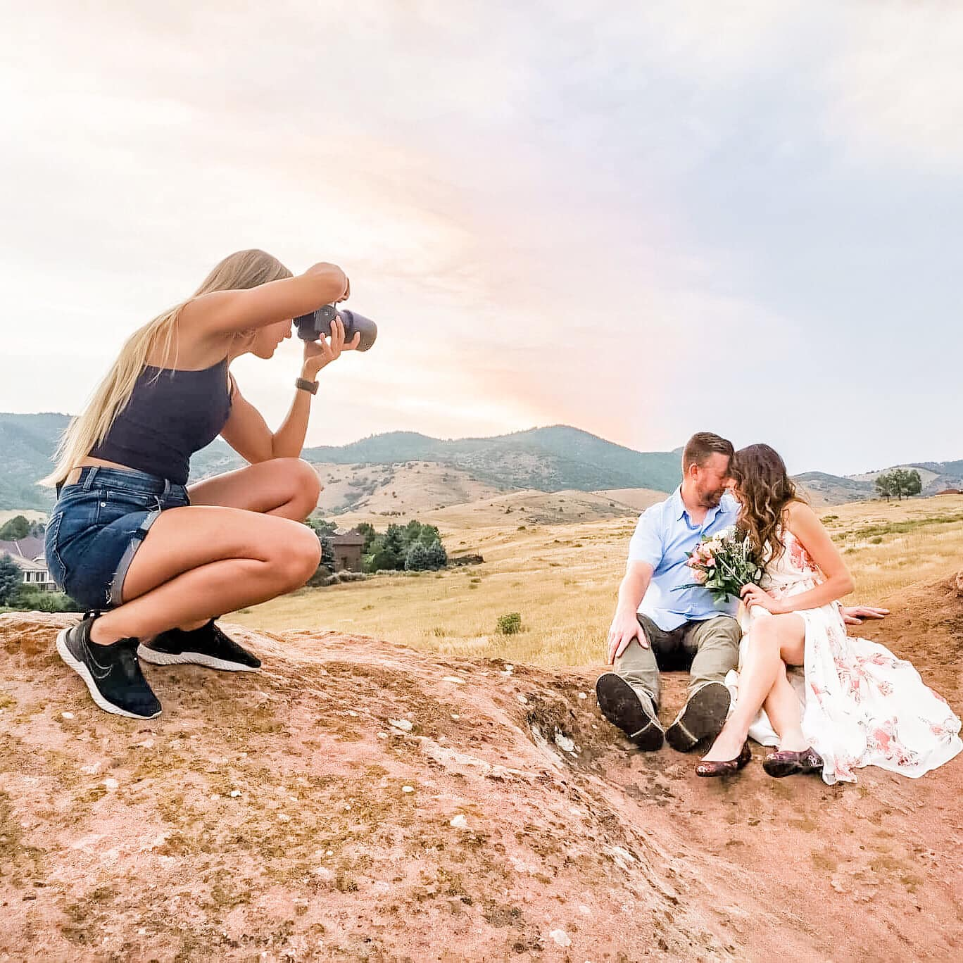 A woman crouching on a rock taking photos of a couple sitting on the ground in a field. The couple is holding hands, and the woman is holding a bouquet, with hills in the background.