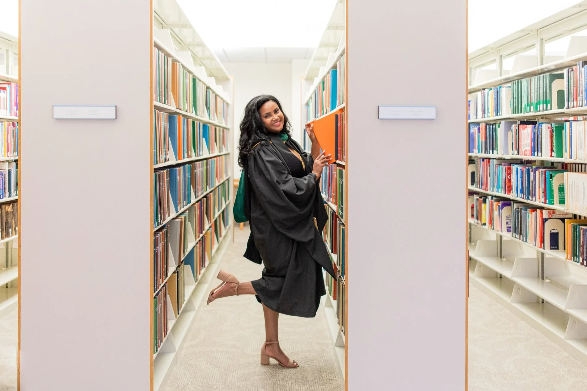 A woman in a graduation gown kneeling between bookshelves in a library, holding an orange book and smiling.