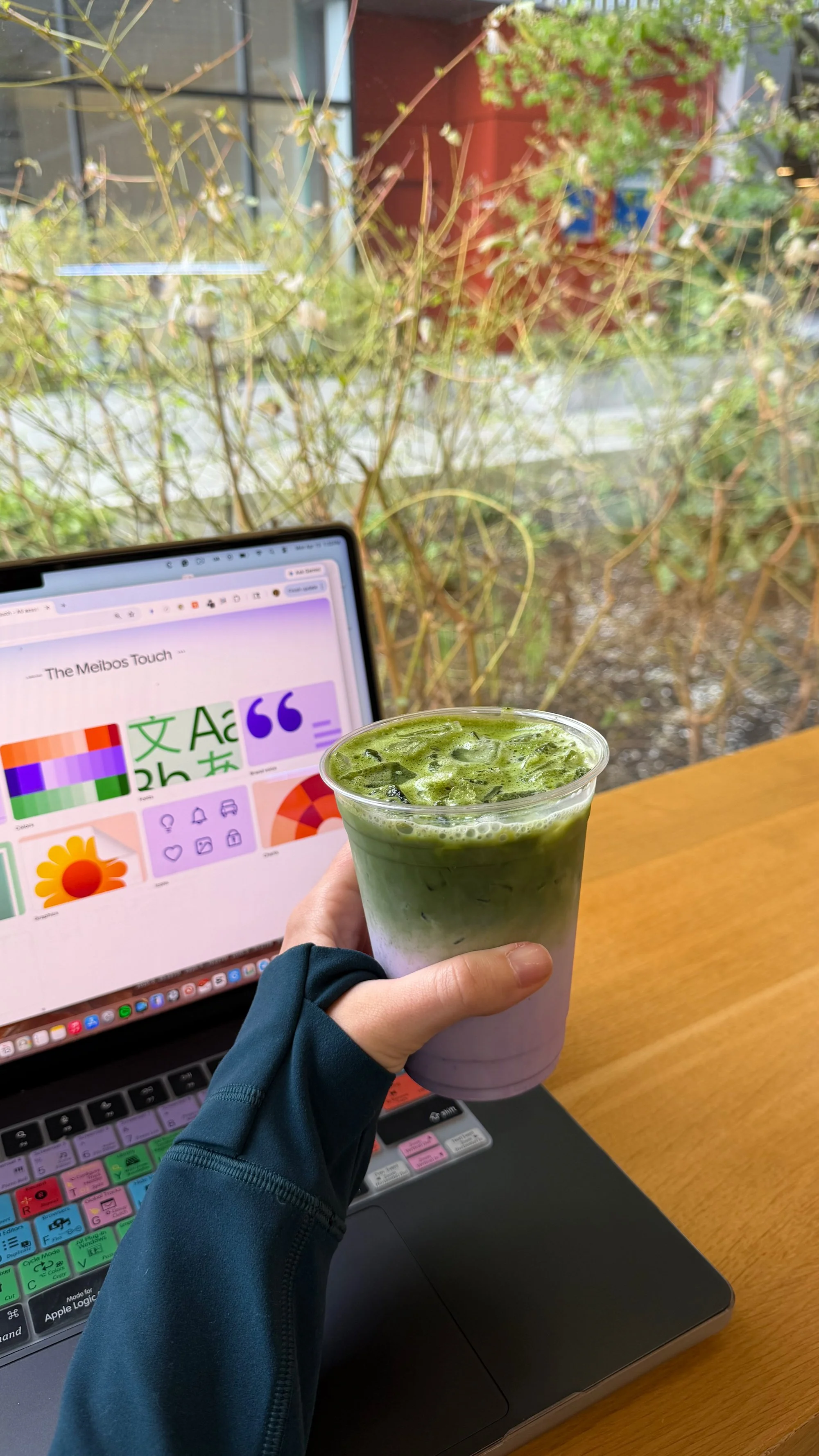 A person holding a green matcha latte. They are sitting at a wooden table with a laptop open to a color palette and graphic design website. In the background, there are large windows with plants outside.
