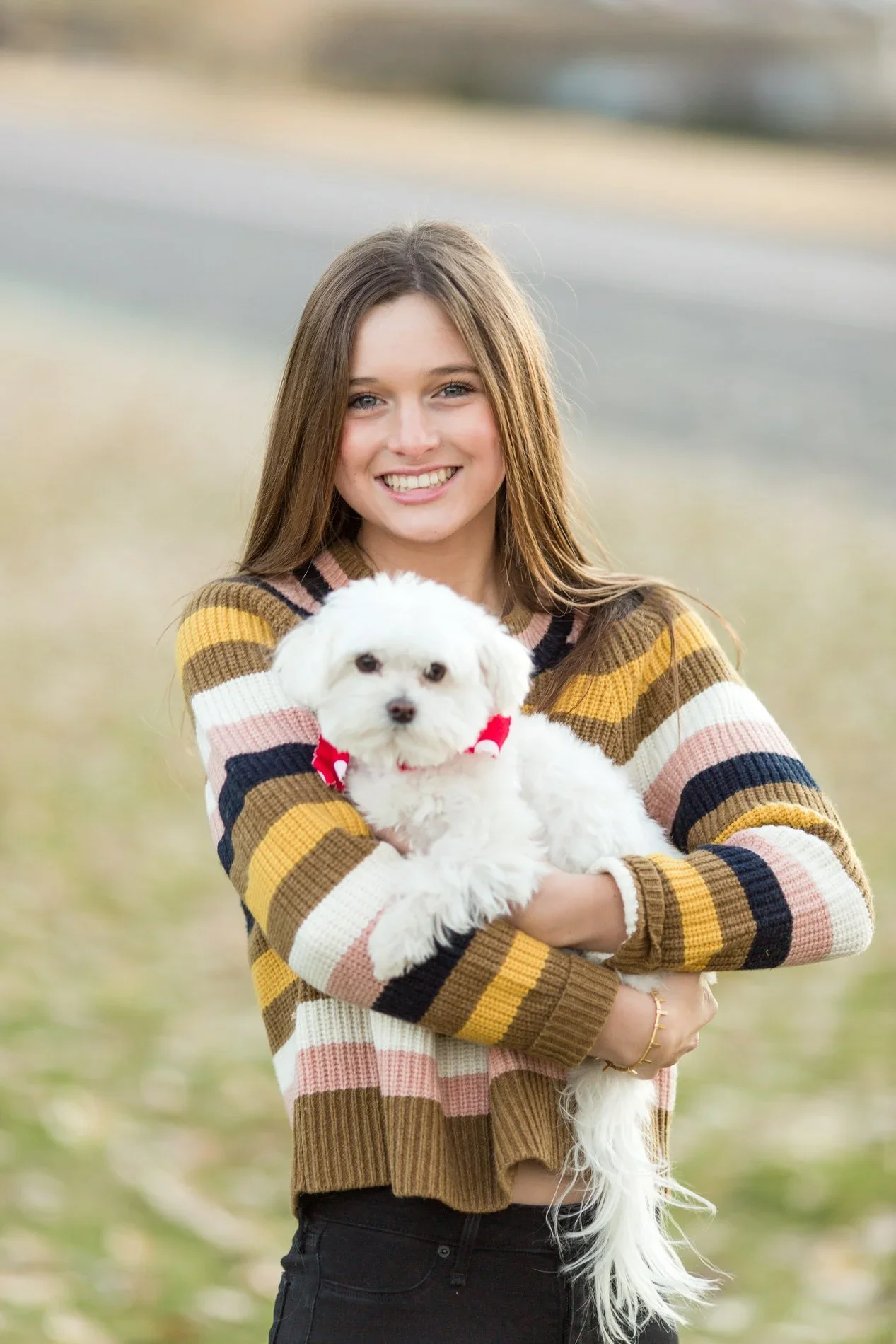 A young woman with long brown hair smiling and holding a small white dog outdoors in a park.