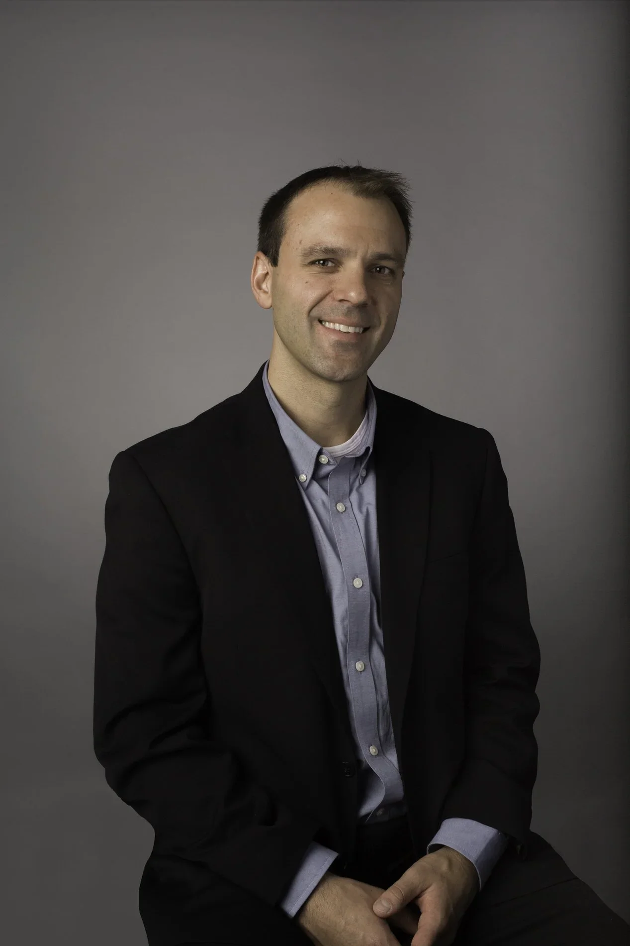 Young man in a black suit and light blue shirt sitting against a plain gray background, smiling at the camera.