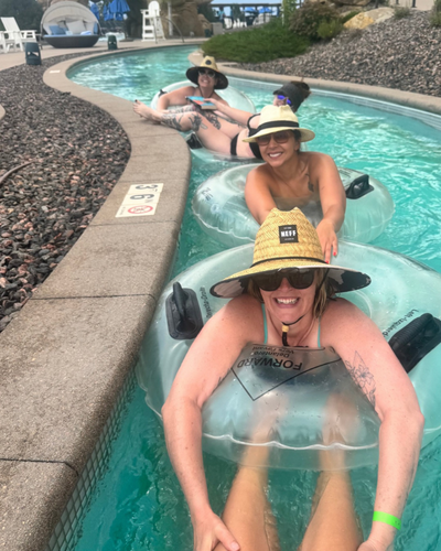 Three women in inflatable tubes relaxing in a lazy river at a water park, all wearing wide-brimmed hats and sunglasses.
