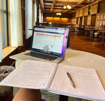 A laptop and open notebooks with a pen are on a white table. The laptop screen displays a webpage, and the notebooks contain handwritten notes. The setting is a cozy, wooden-furnished cafe or library with large windows and warm lighting.