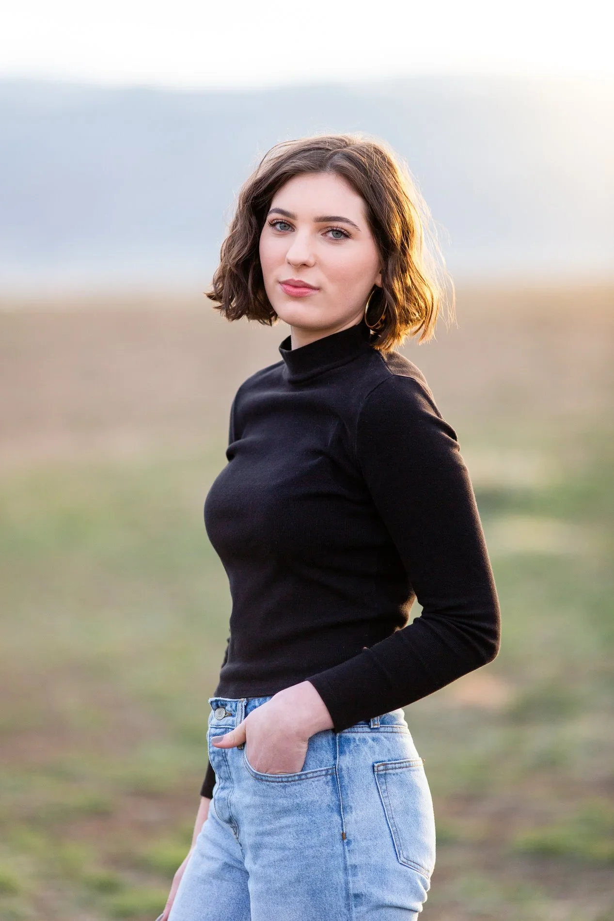 A young woman with shoulder-length brown hair and blue eyes standing outdoors in a field, wearing a black turtleneck top and light blue jeans, with her right hand in her pocket.